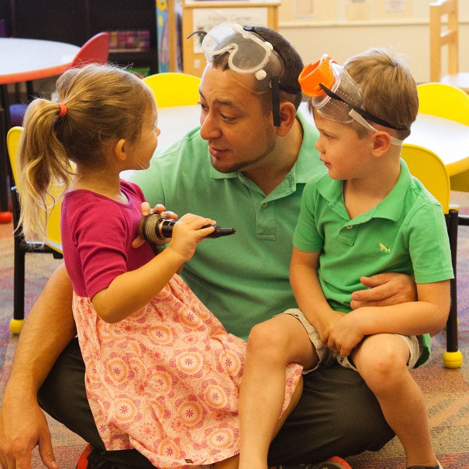 Person with two children wearing safety goggles in a classroom