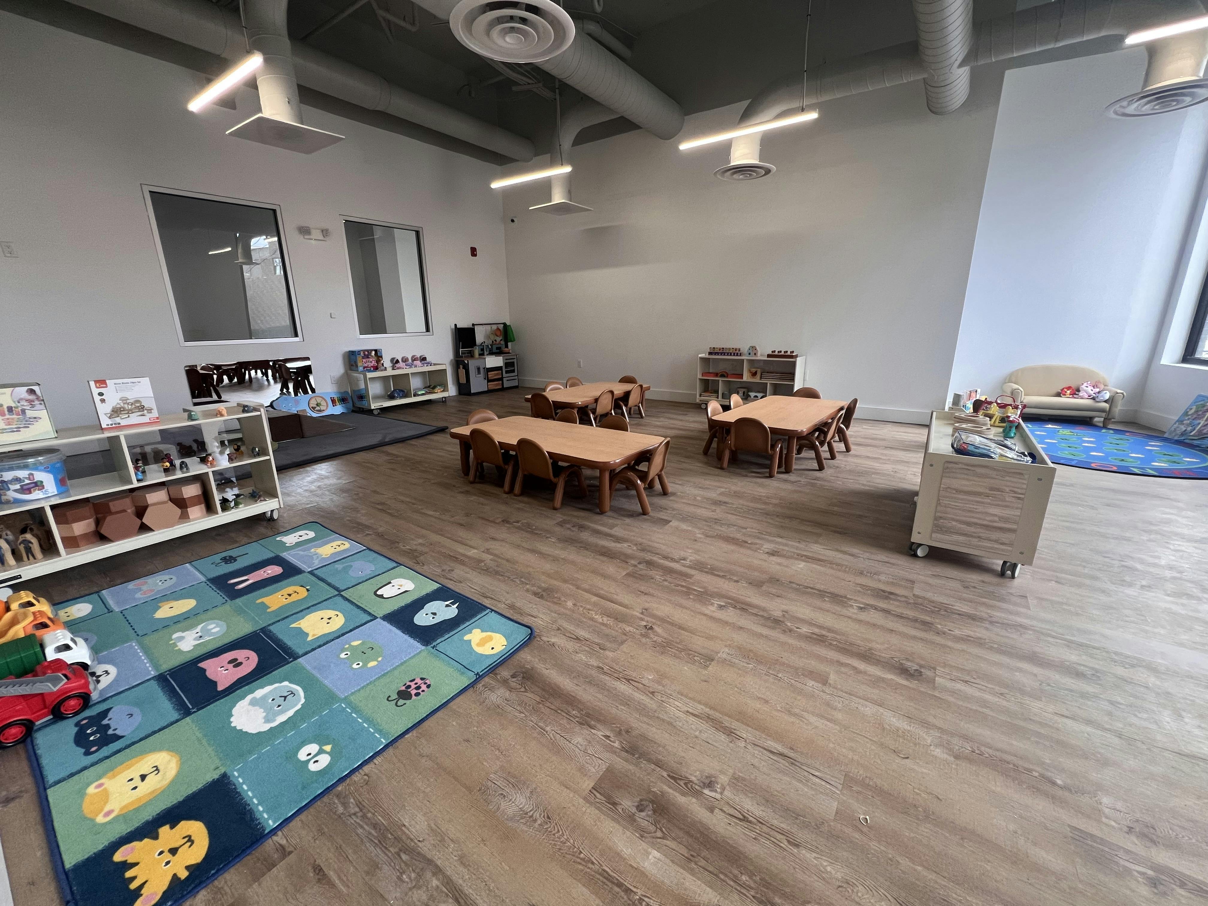 Empty classroom with small wooden tables and chairs, play mats, and toy shelves