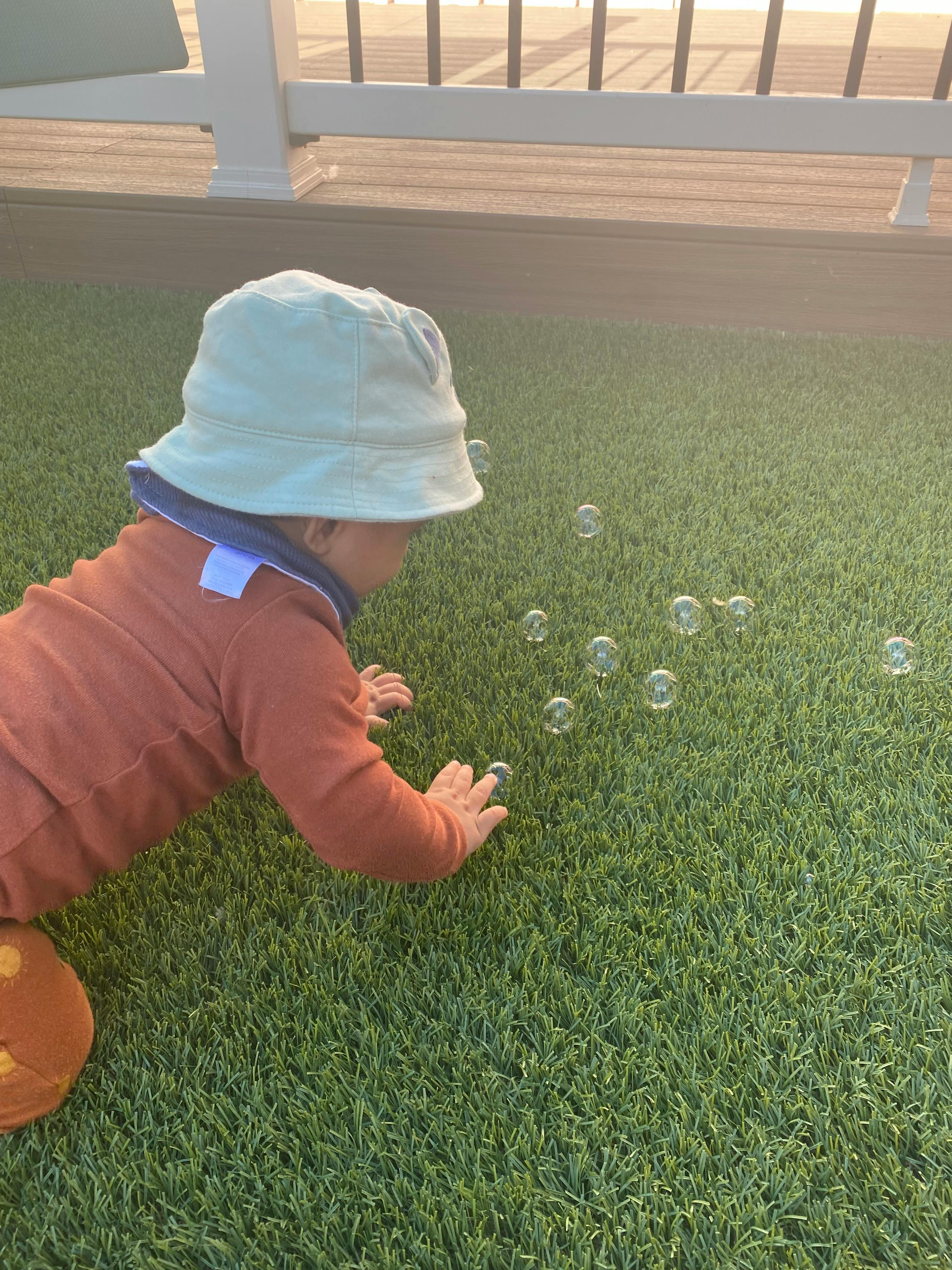 Child in a hat crawling on grass toward bubbles
