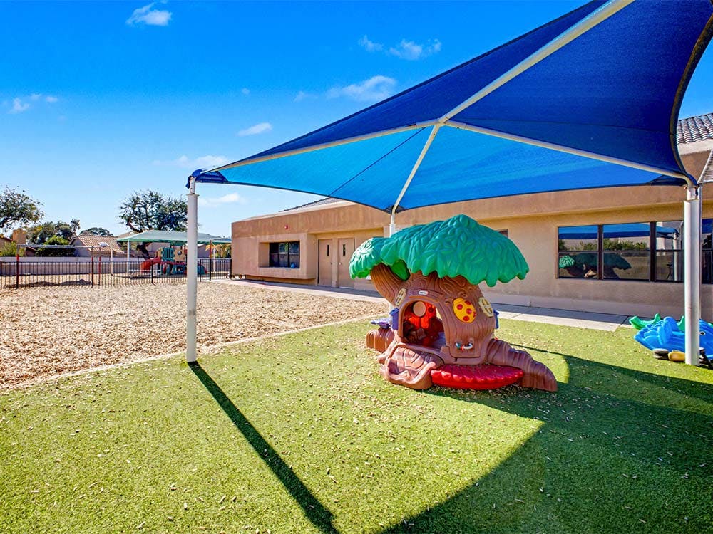 Playground with artificial grass, wood chips, and a play treehouse under a blue shade
