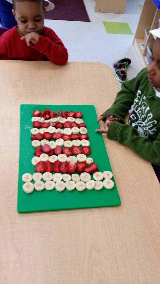 Two children at a table with a green cutting board of strawberries and bananas
