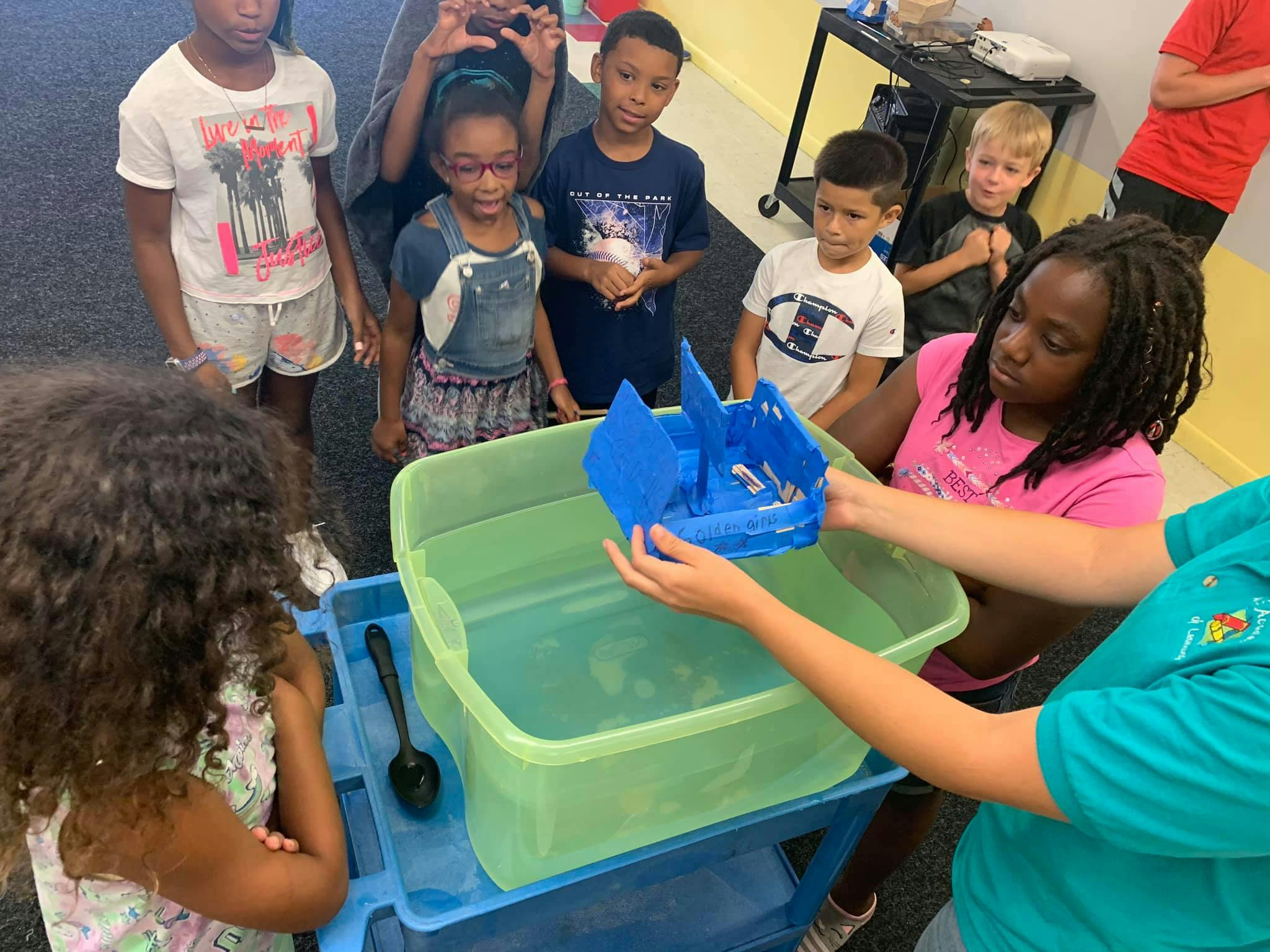 Children gathered around a bin of water, one person holding a blue toy boat