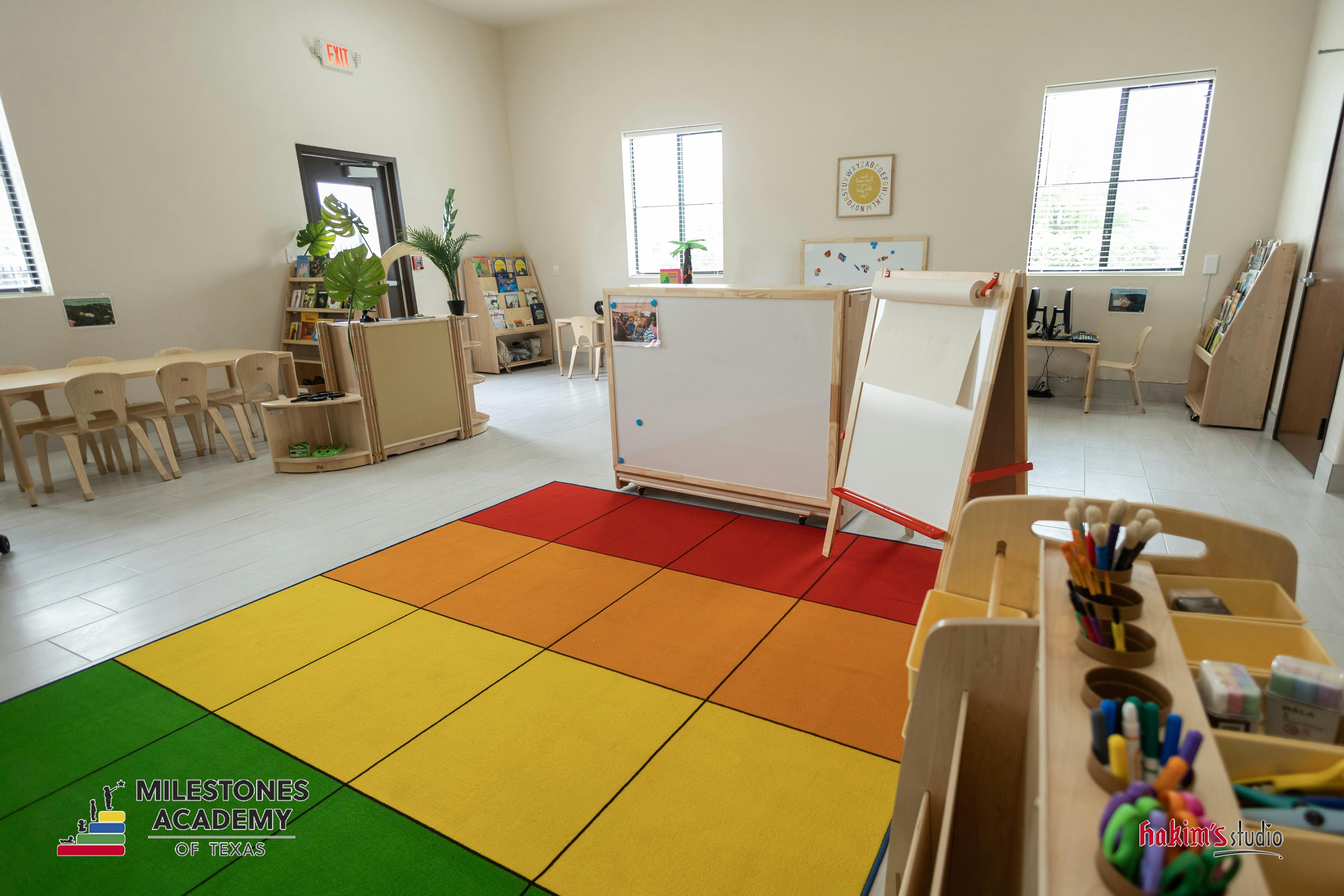 An empty, brightly lit classroom with child-sized tables and chairs, easels, and a colorful rug