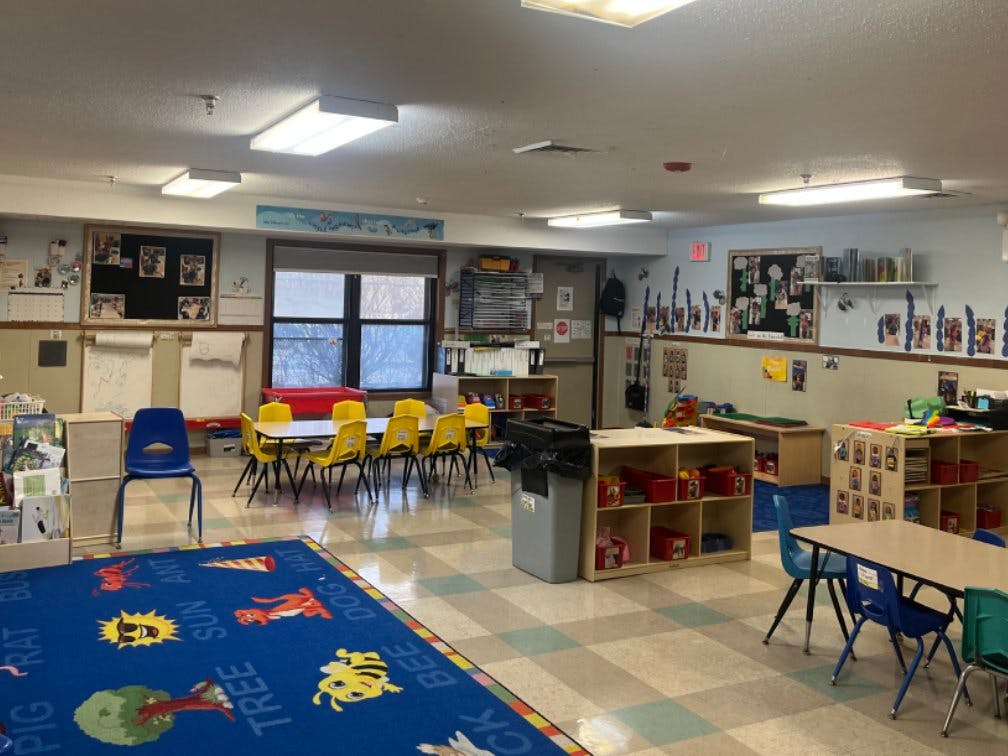 Empty classroom with small tables and chairs, cubbies, and a colorful alphabet rug