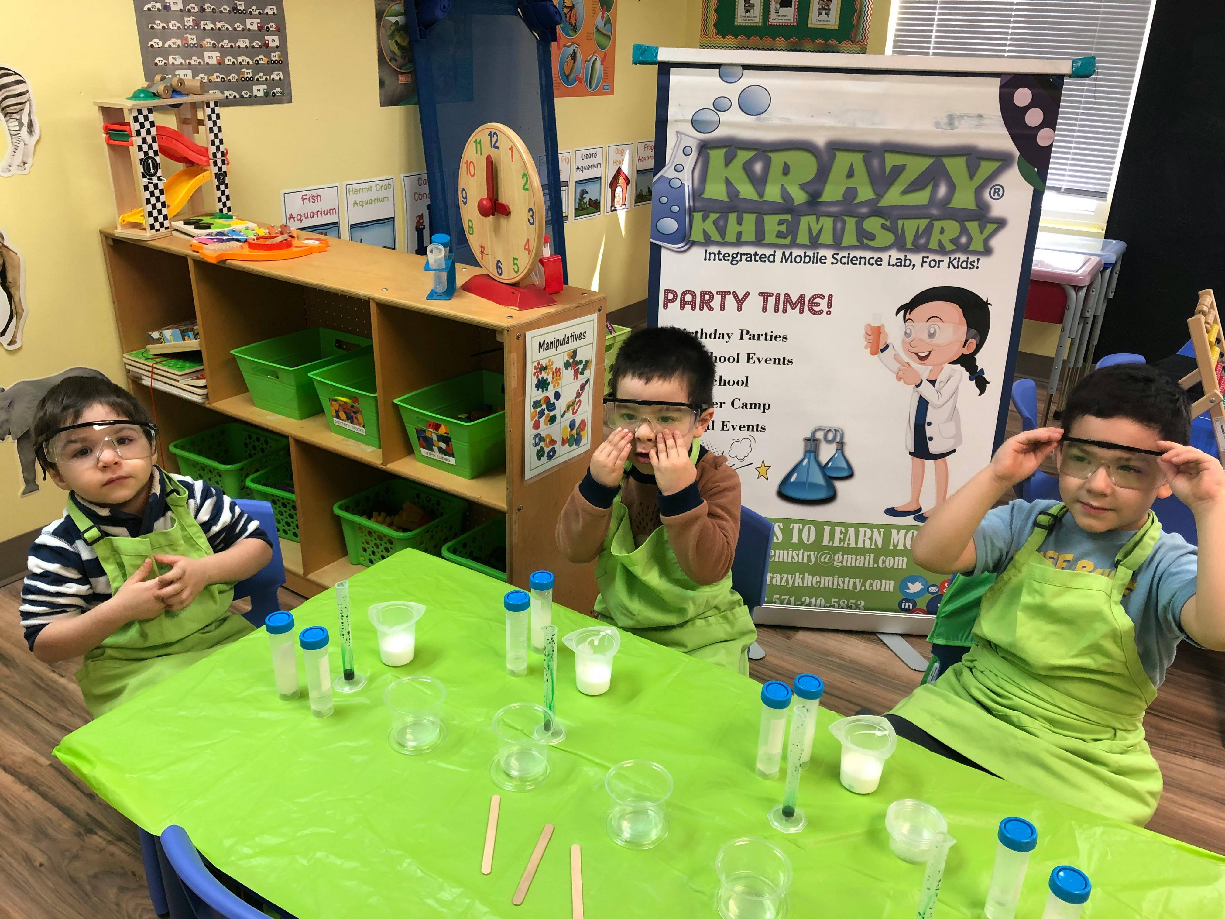 Three children in safety goggles and aprons at a table with beakers