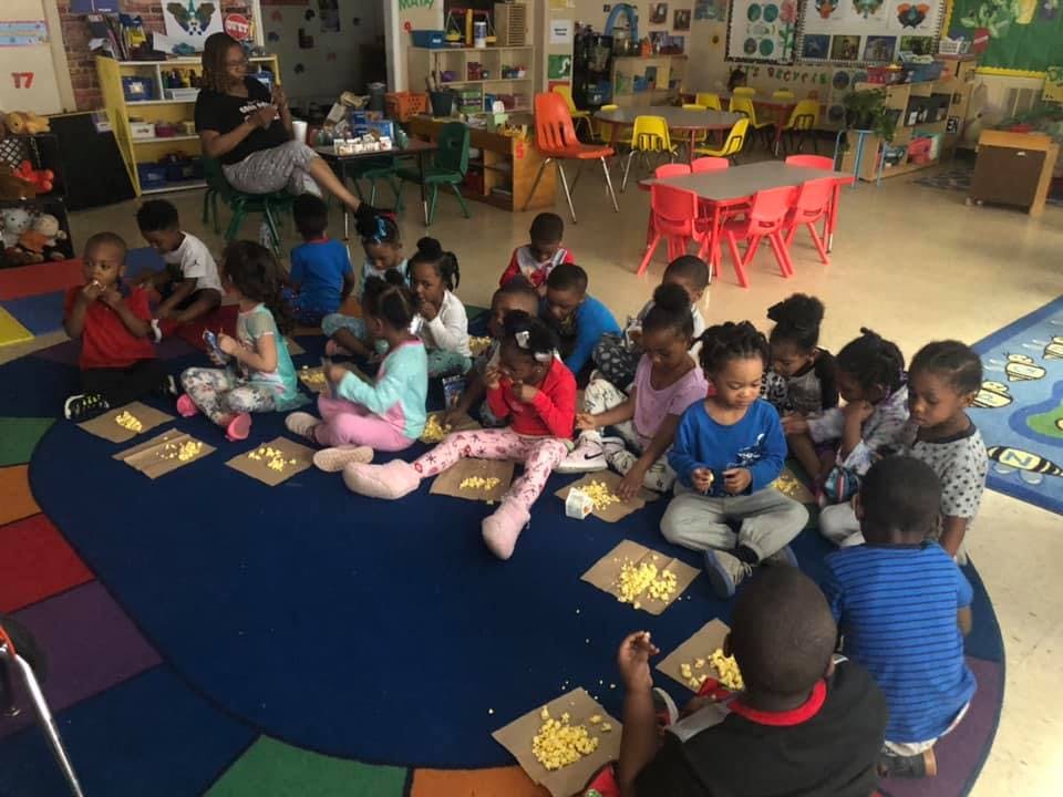 Many children sitting on a rug in a classroom, some with small piles of food
