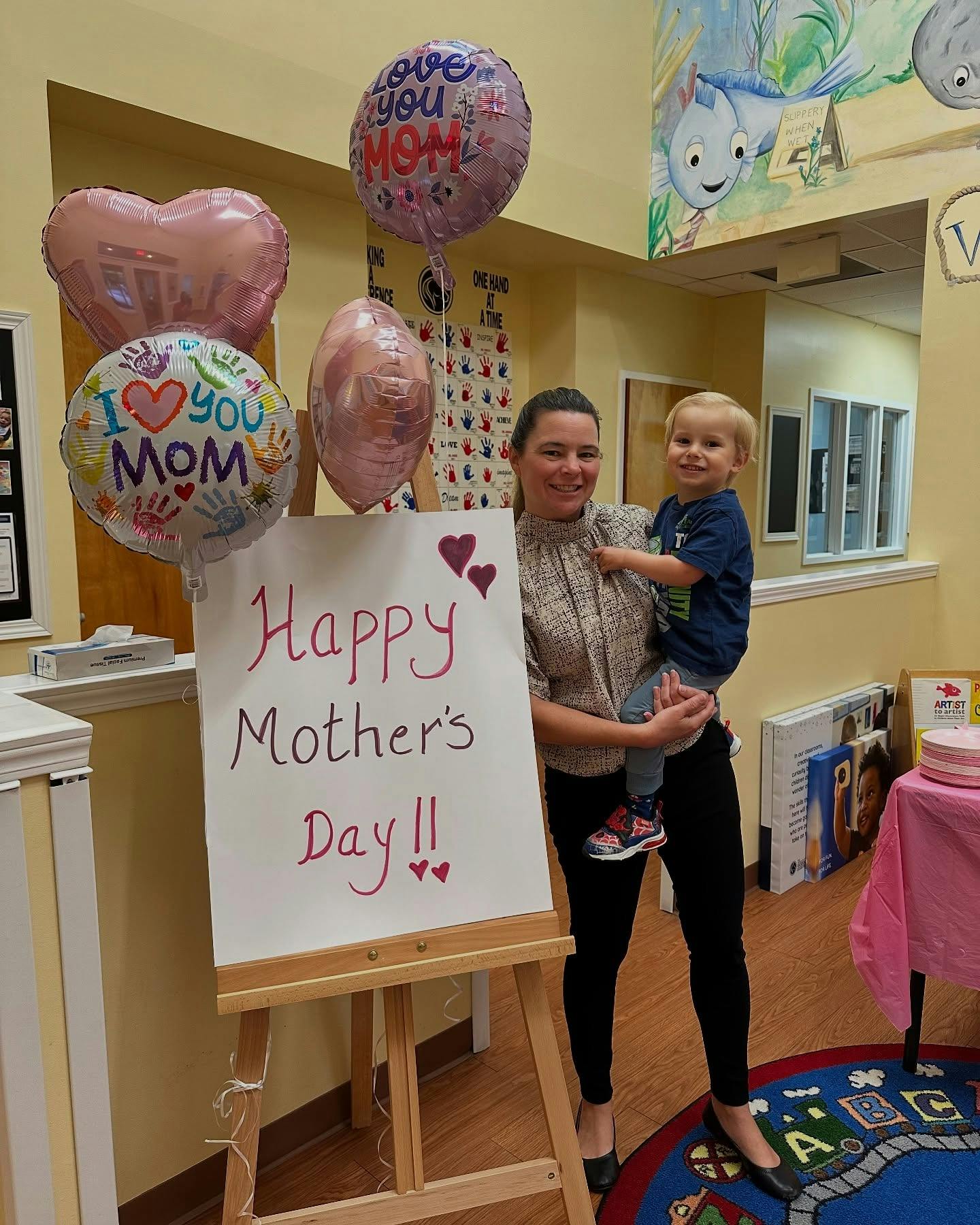Person holding a child next to a "Happy Mother's Day" sign and balloons