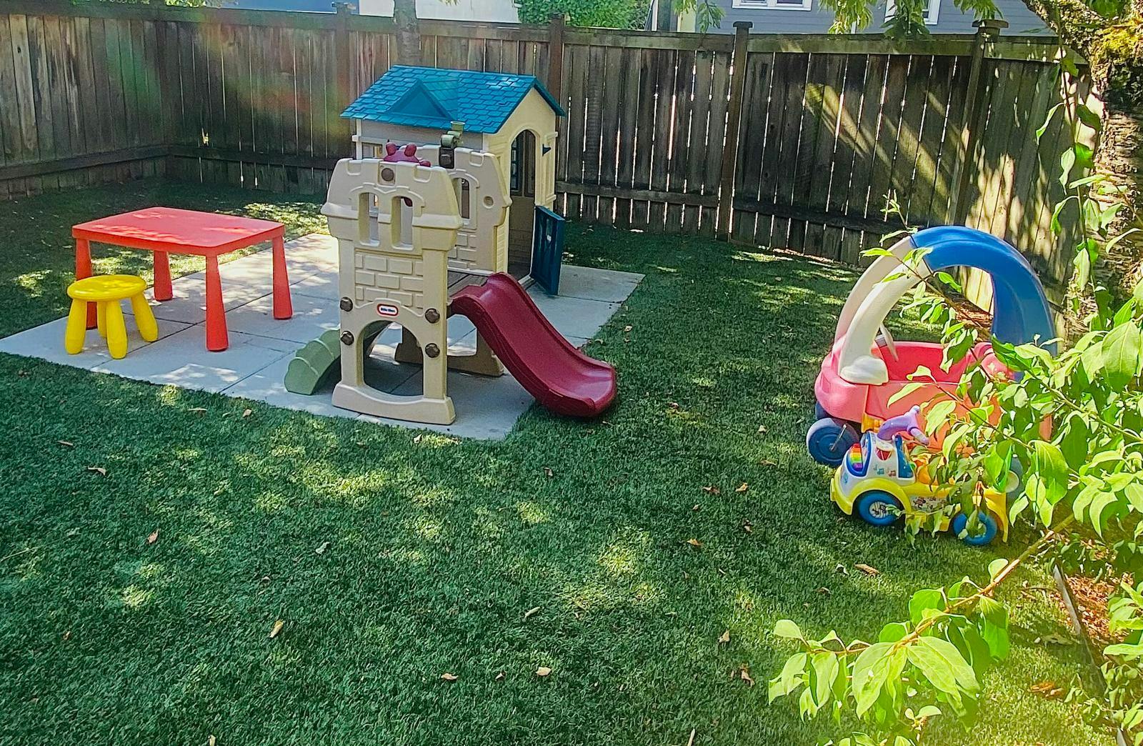 Playhouse with slide, red table, and toy car in a grassy backyard
