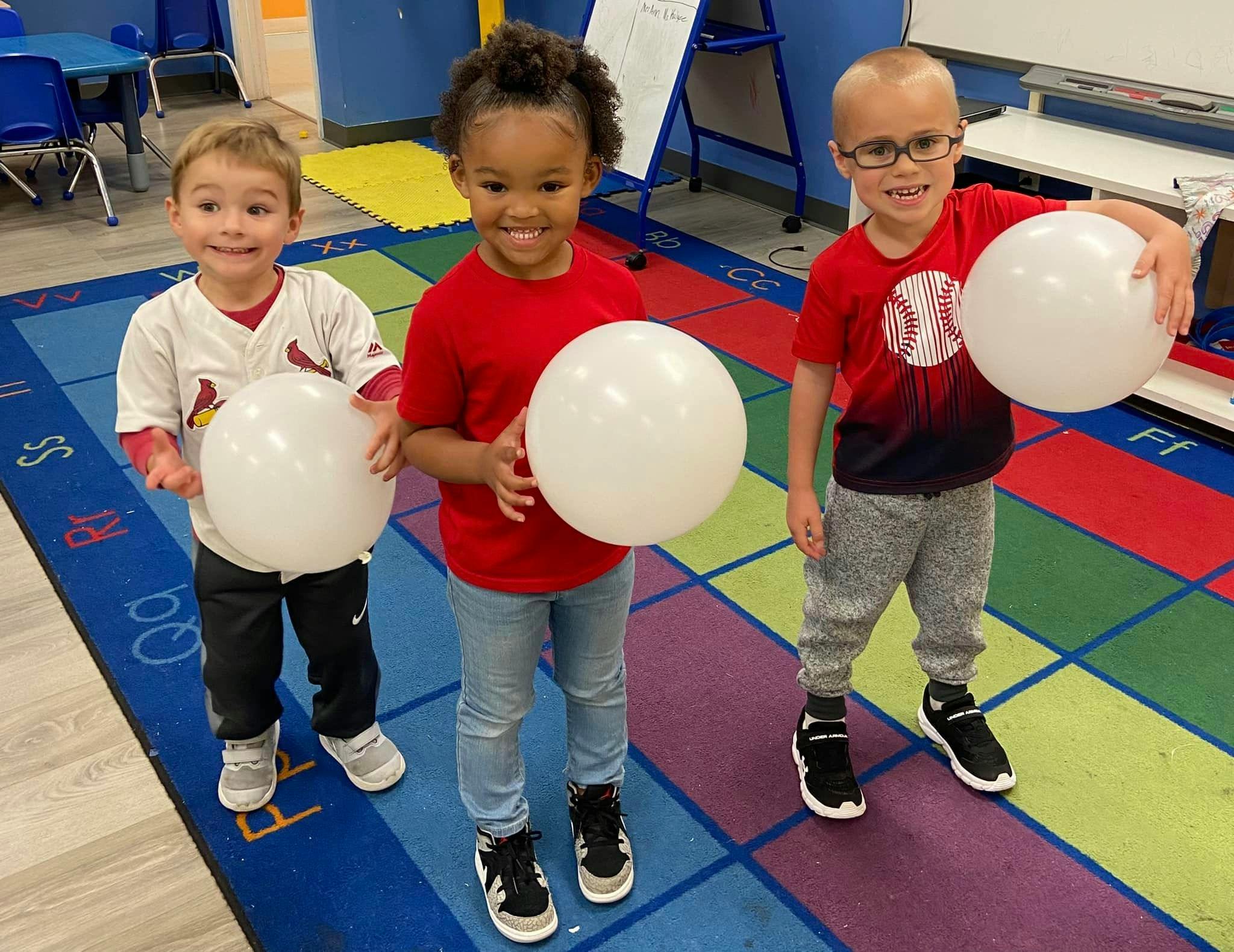 Three children holding white balloons in a classroom