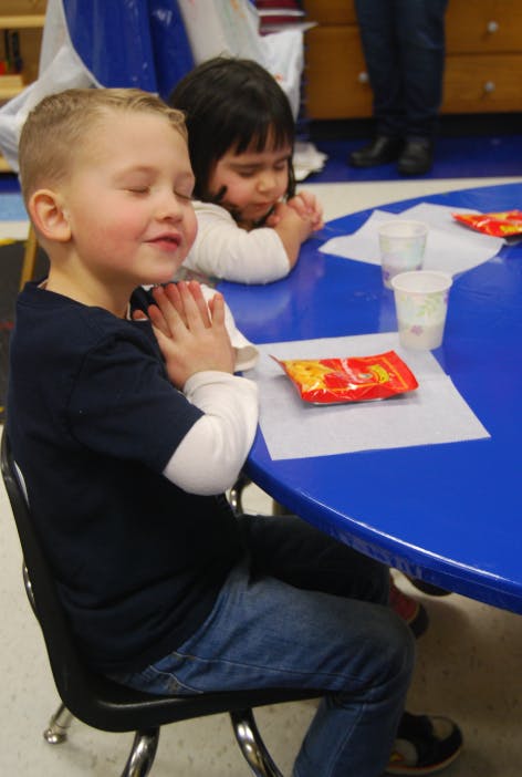 Child with eyes closed and hands together at a table with another child