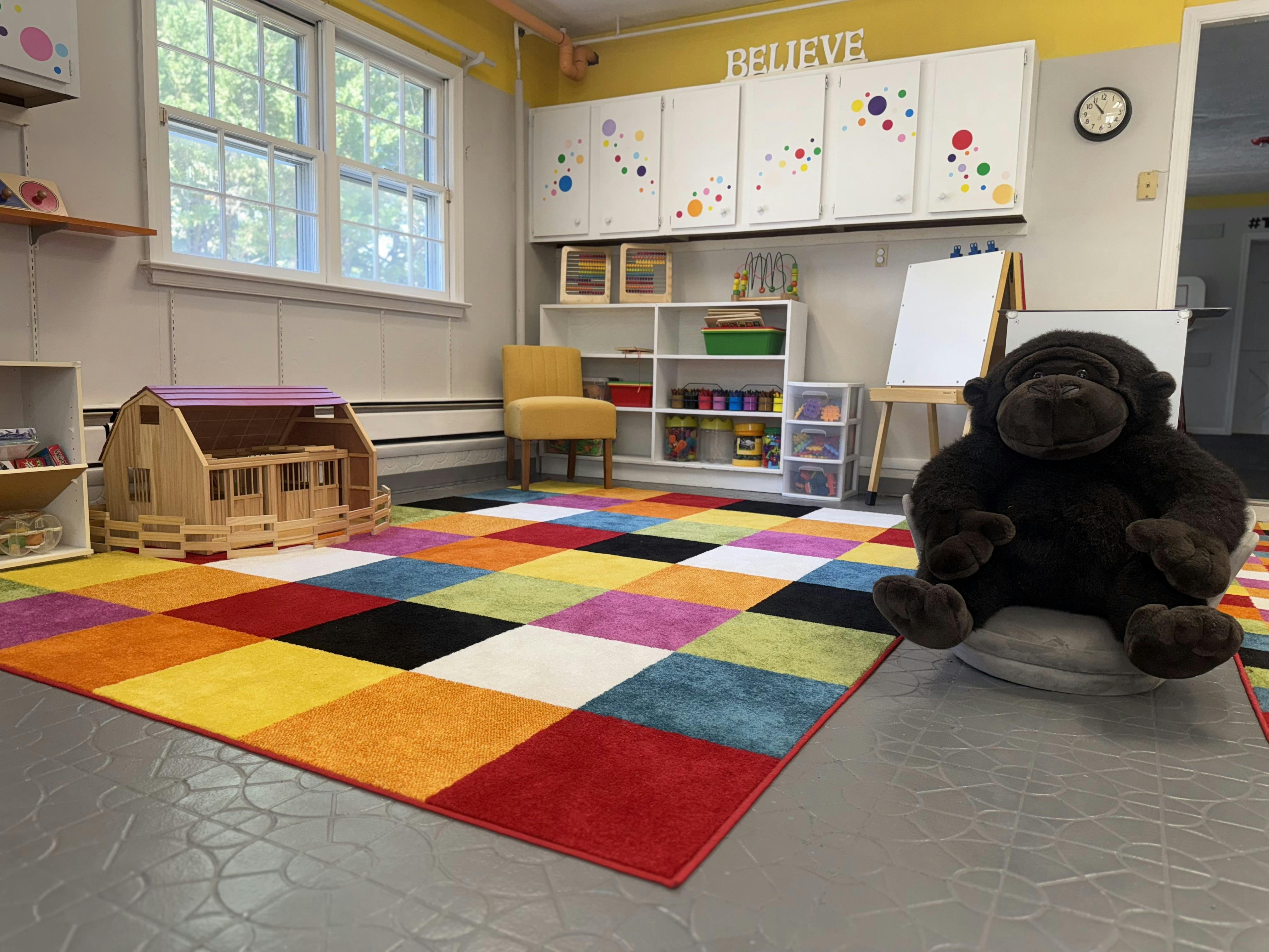 Classroom with colorful rug, toy barn, white cabinets, and large stuffed gorilla