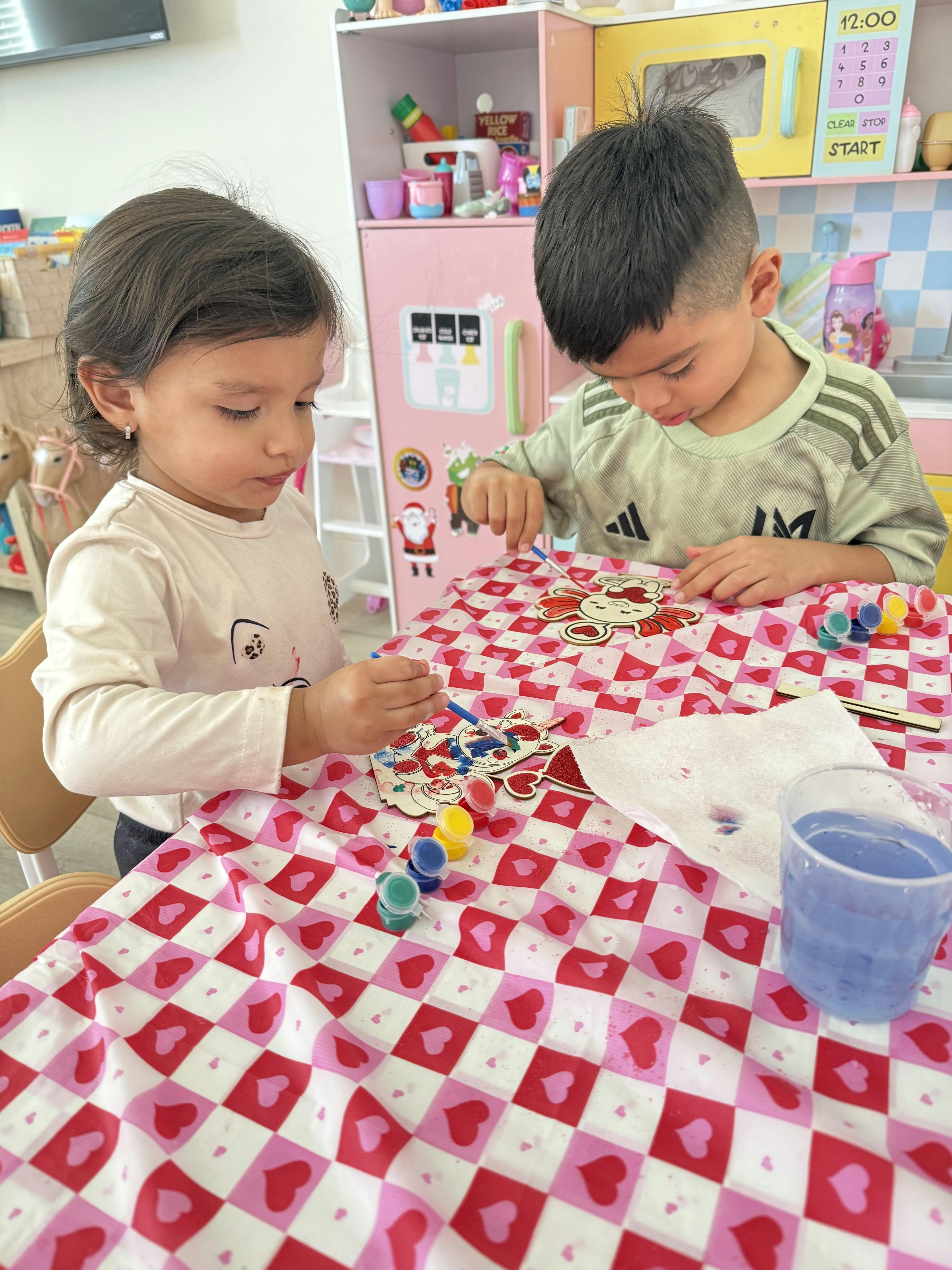 Two children painting wooden cutouts at a table in a play area