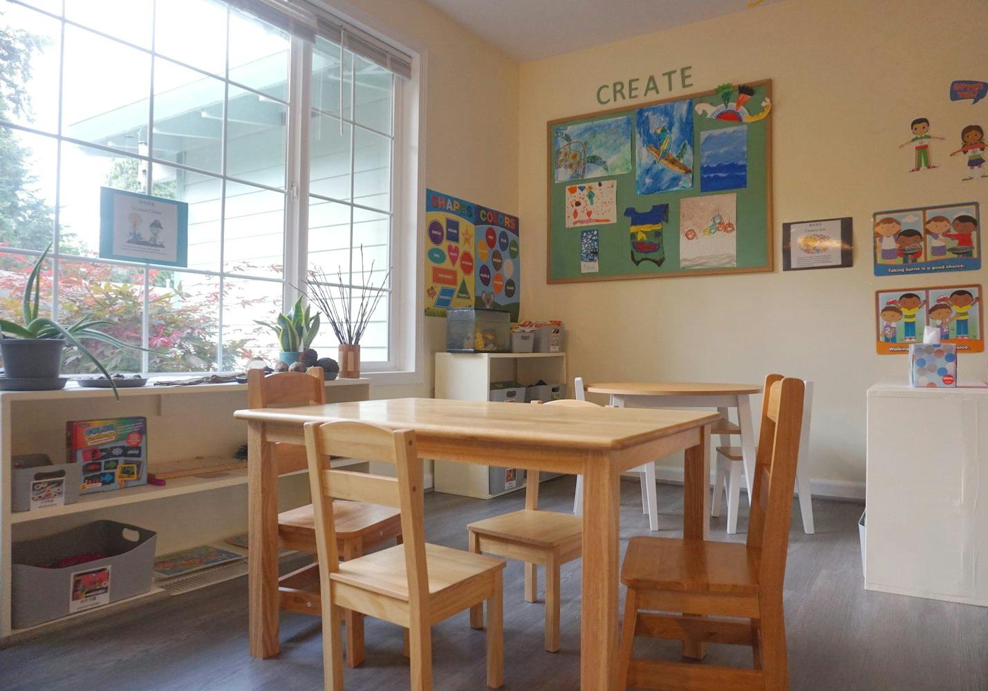 Empty classroom with small wooden tables and chairs, bulletin board with children's art