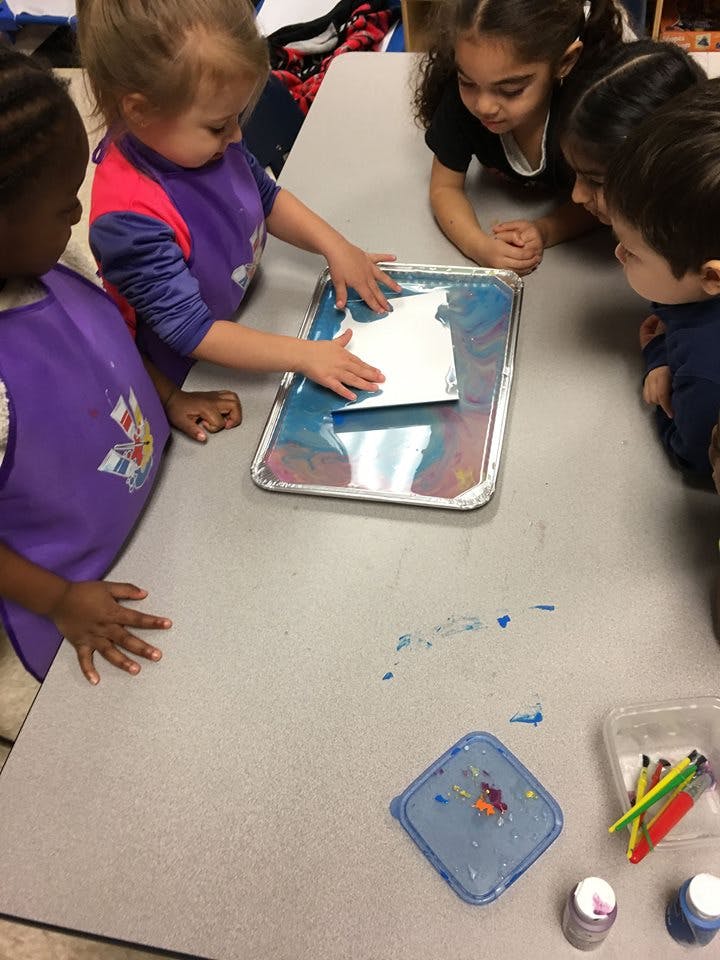 Children gathered around a table, one child pressing paper into a tray of liquid