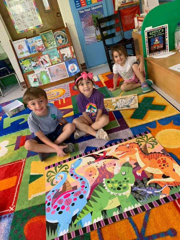 Three children sitting on a colorful rug with a dinosaur puzzle