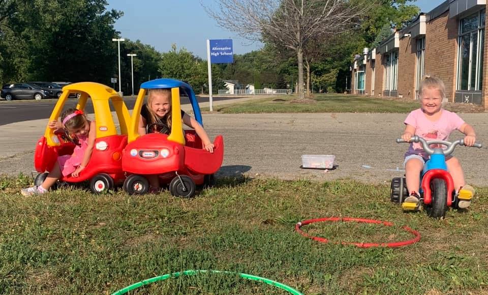 Three children playing with toy cars and a tricycle outdoors