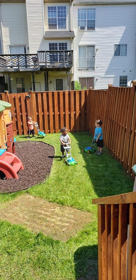 Three children playing with toy lawnmowers in a fenced yard