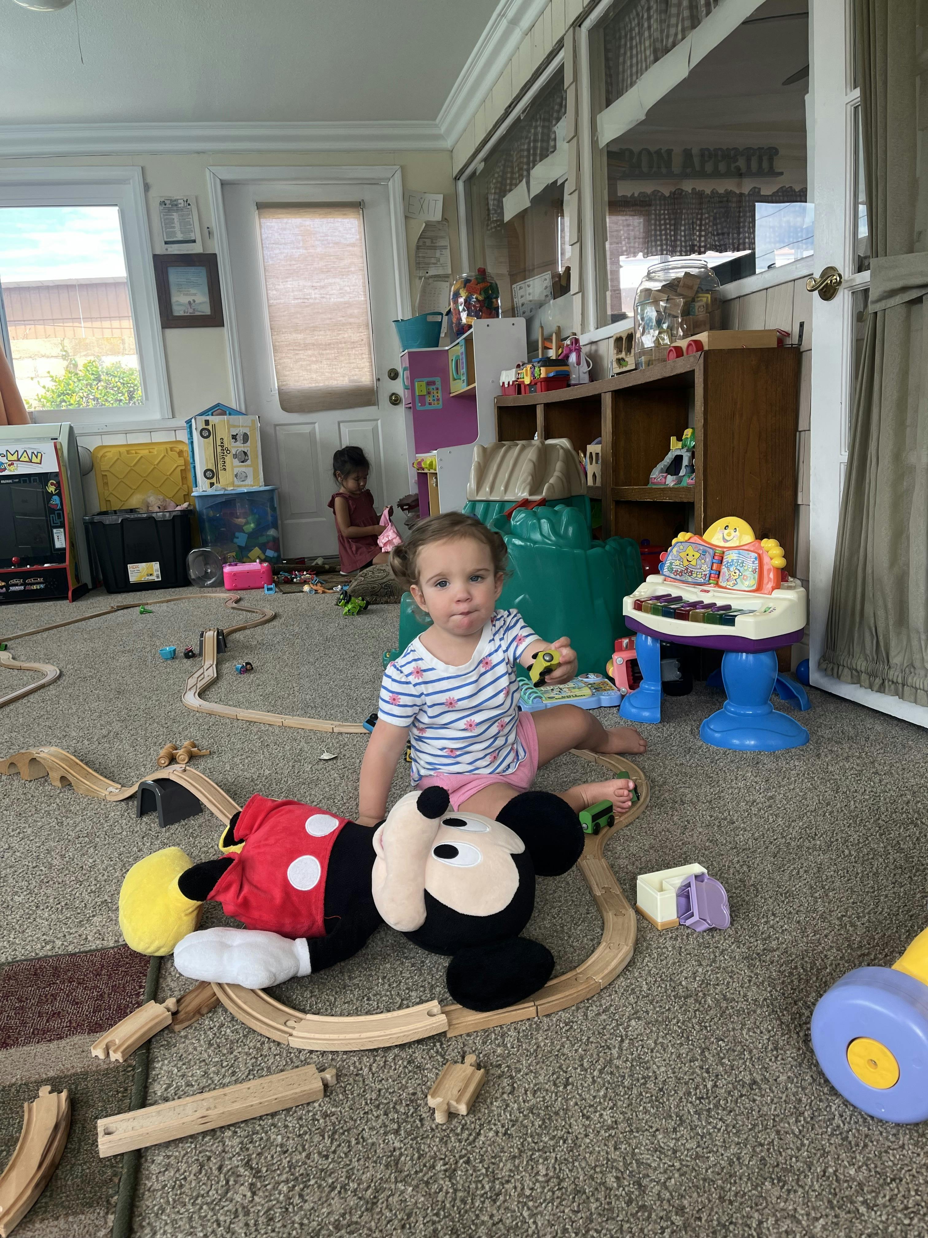 Child sitting on a carpeted floor surrounded by toy train tracks and a plush toy