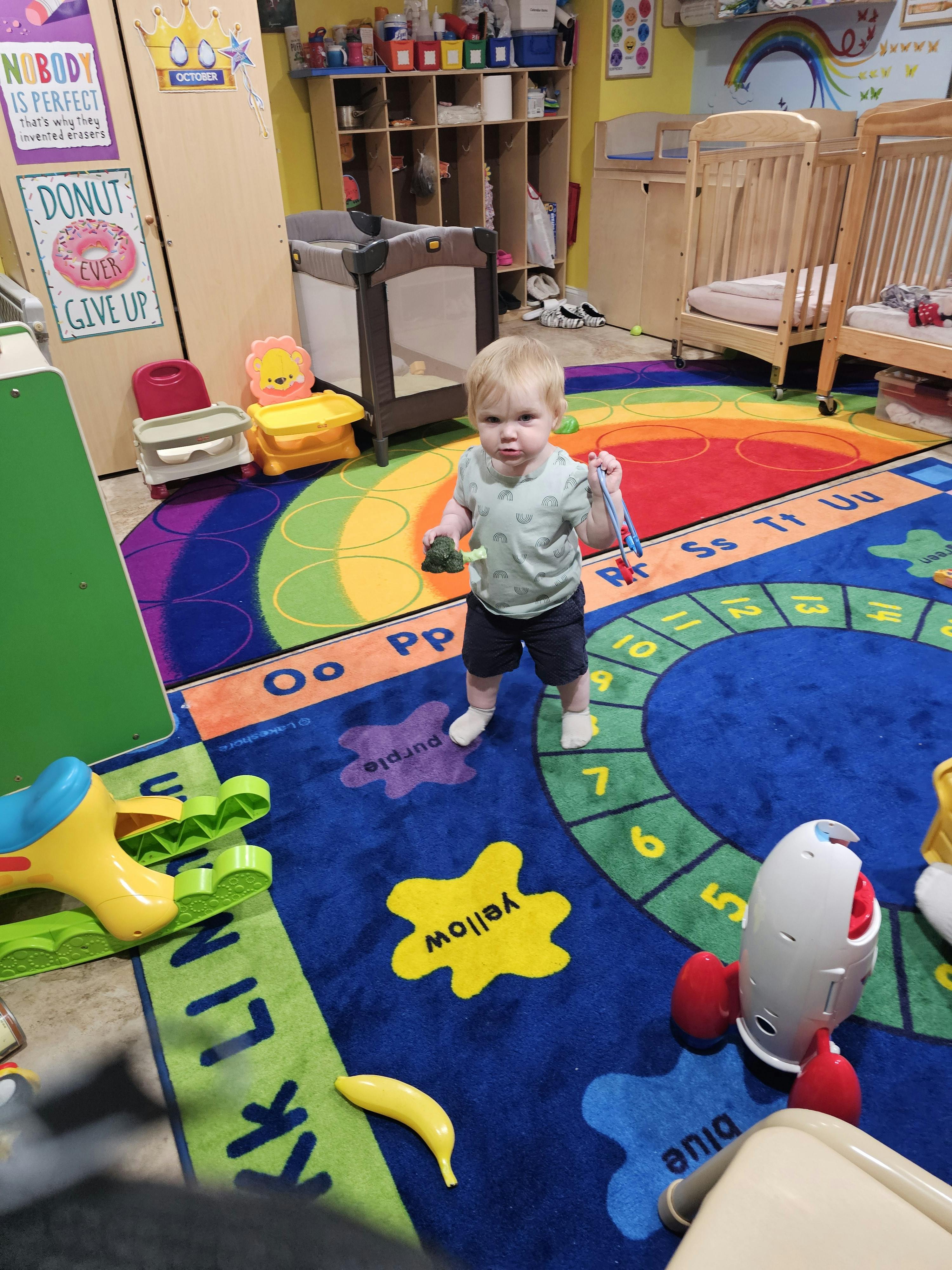 Child holding toy broccoli and tongs on a colorful alphabet rug