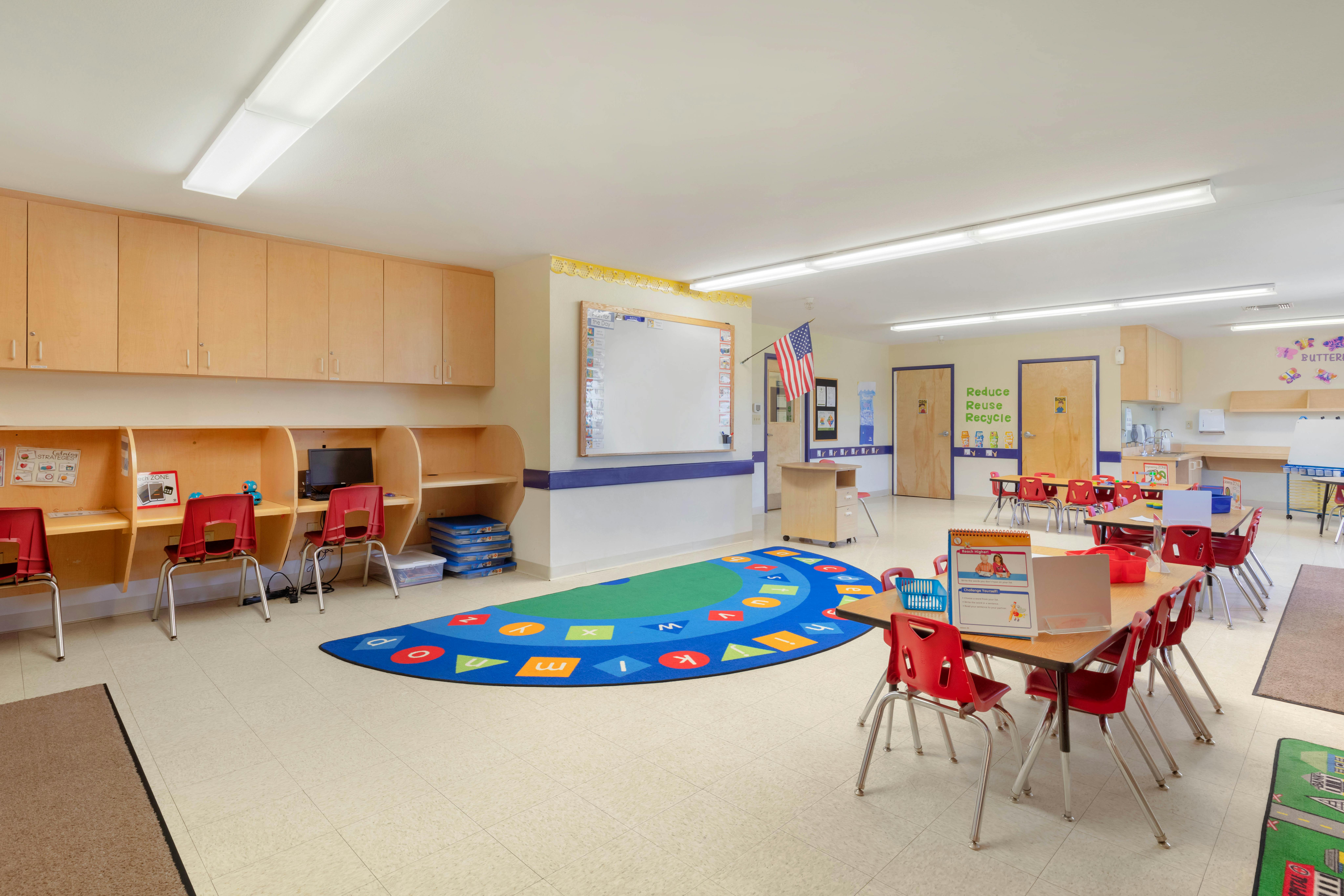 Empty classroom with small red chairs, wooden desks, and an alphabet rug