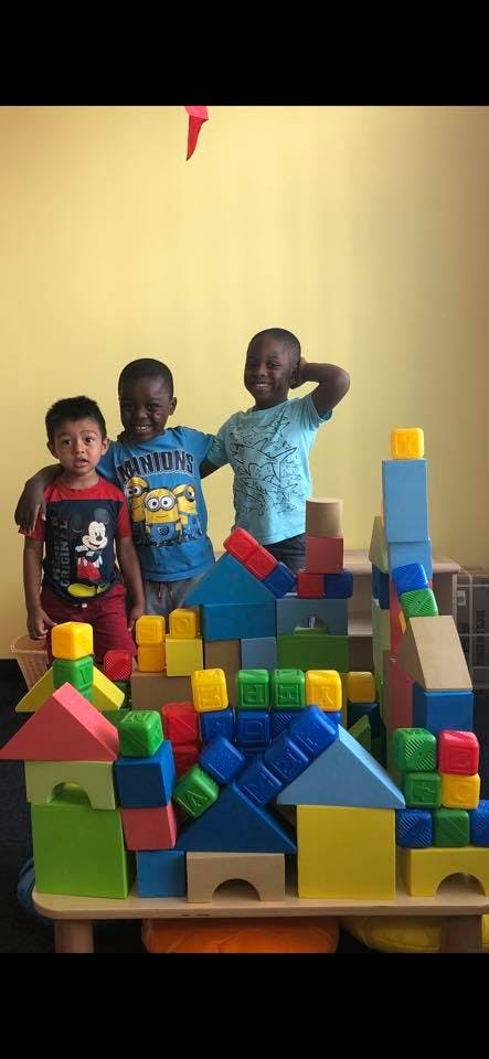 Three children standing behind a large structure made of colorful building blocks