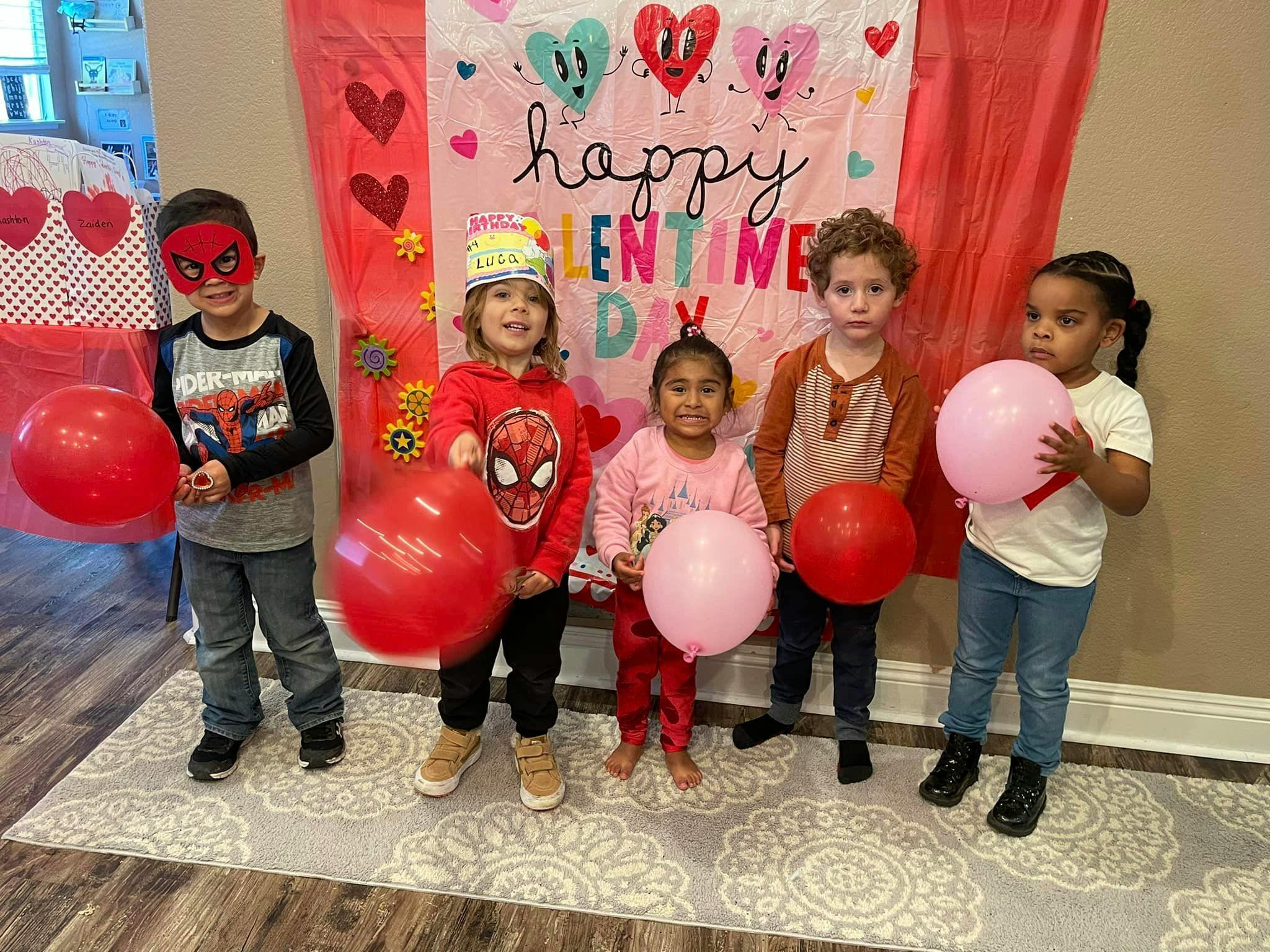 Five children holding balloons in front of a "Happy Valentine's Day" banner
