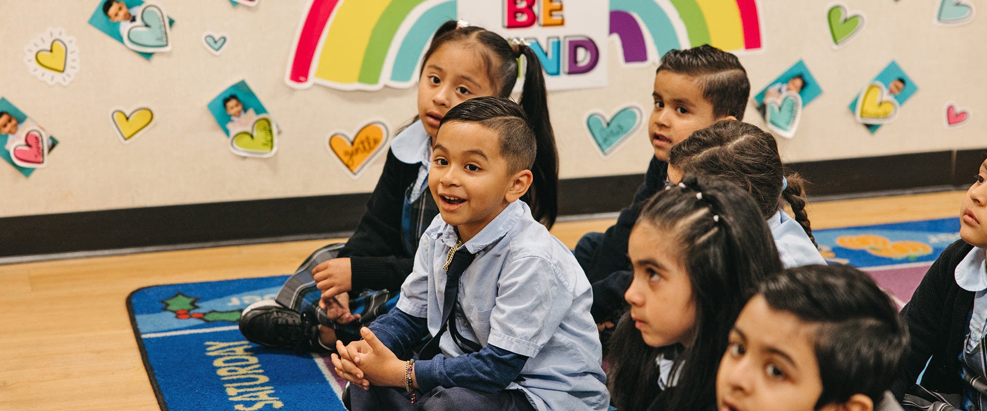Children sitting on a rug in a classroom with Be Kind sign in background