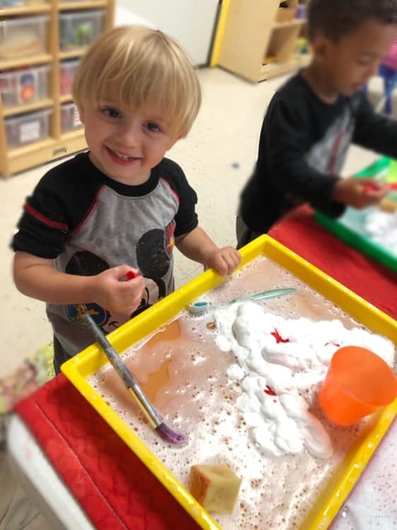 Child holding a red object over a yellow tray with soapy water and a paintbrush
