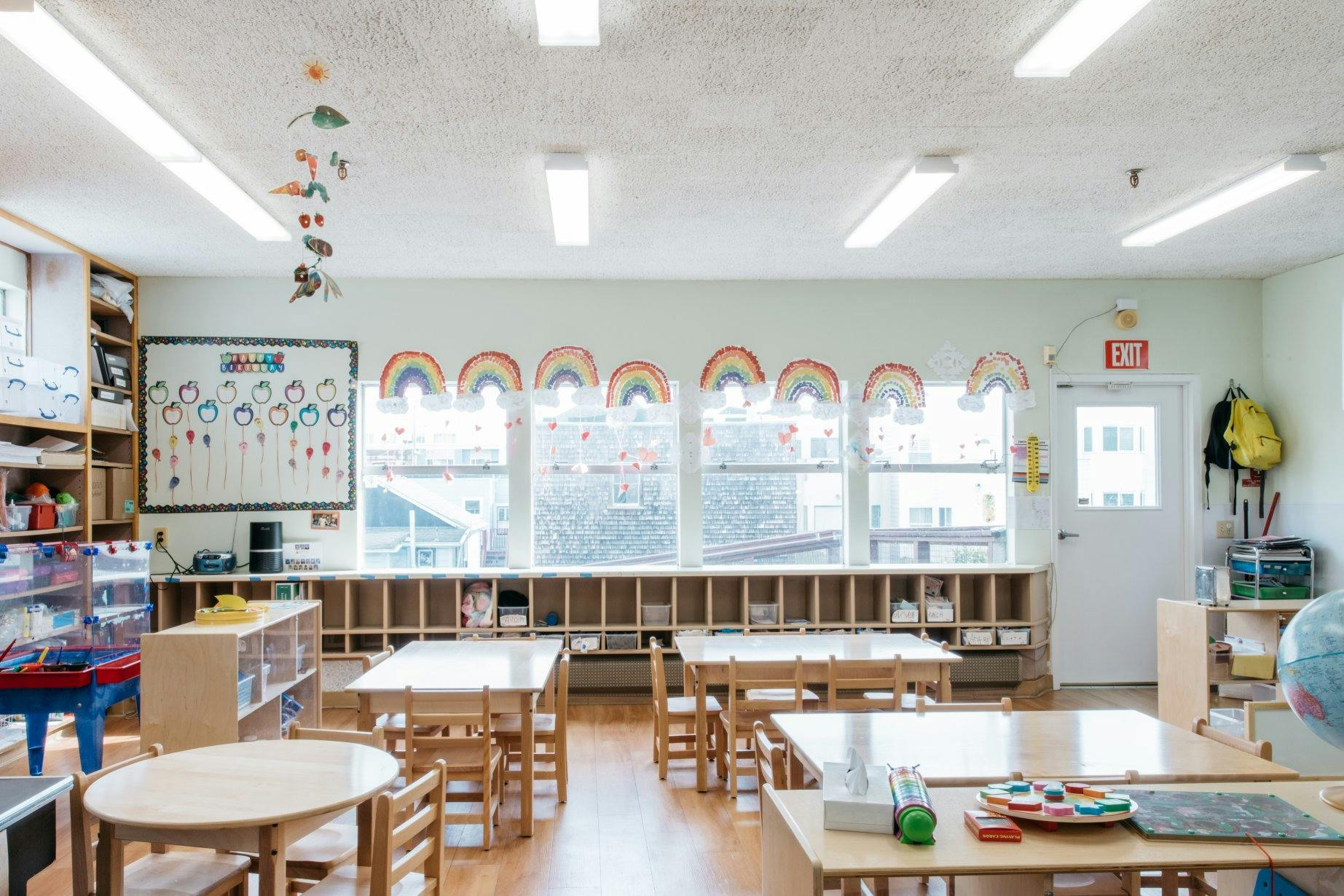 Empty classroom with small tables and chairs, windows with rainbow decorations
