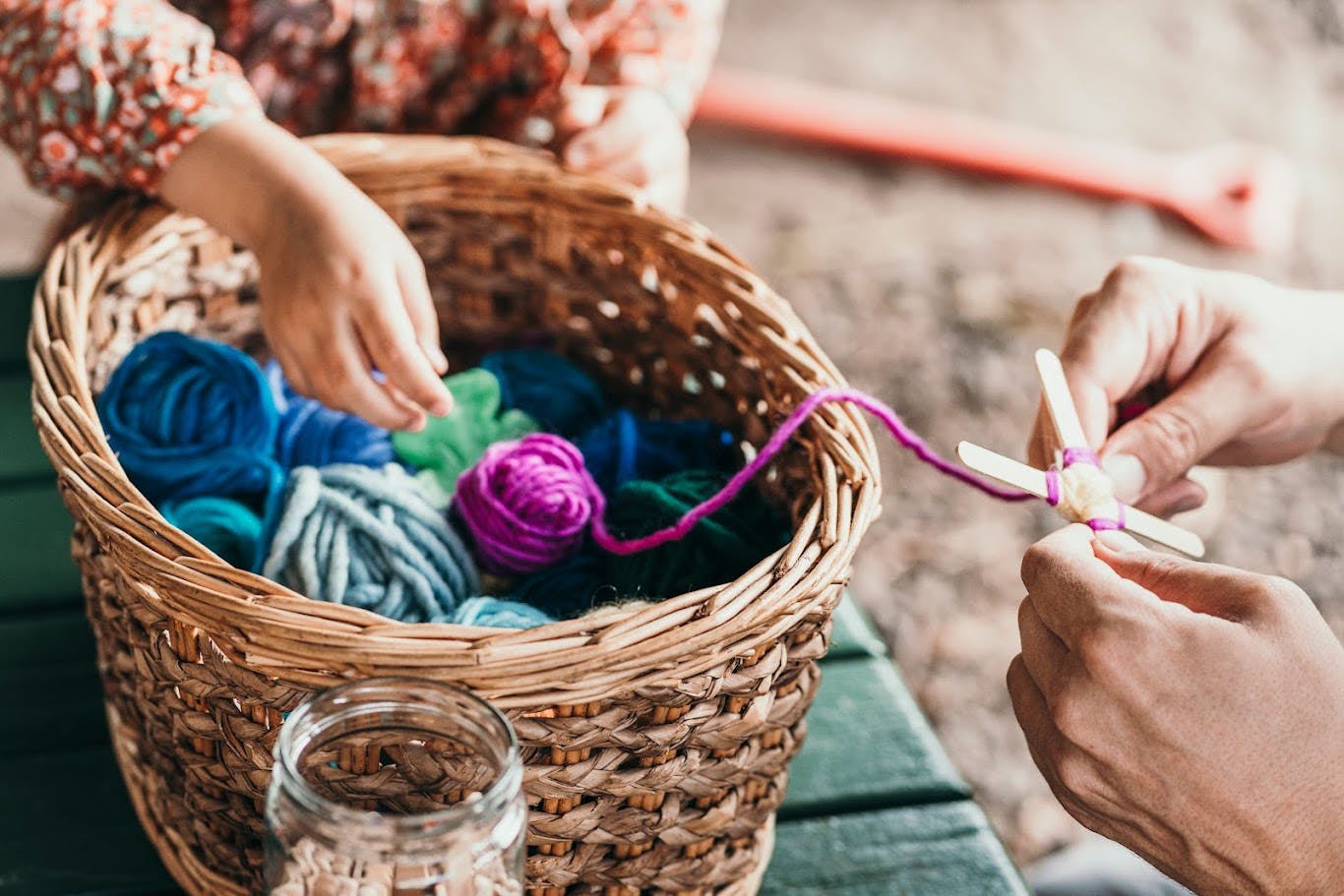 Person's hands working with yarn and craft sticks next to a basket of yarn