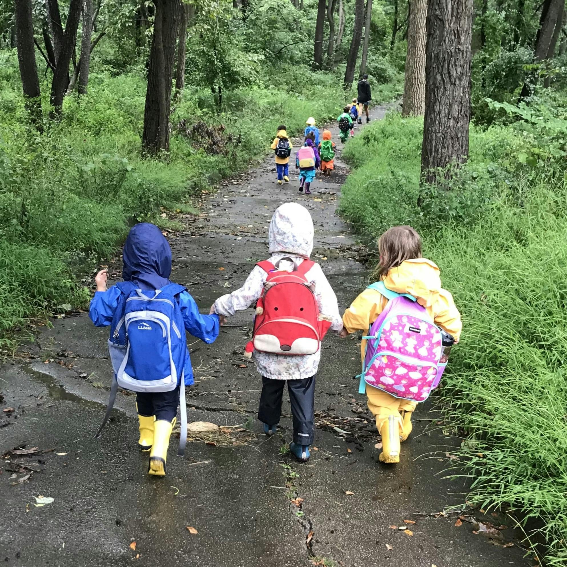 Group of children in rain gear and backpacks walking on a path in a forest