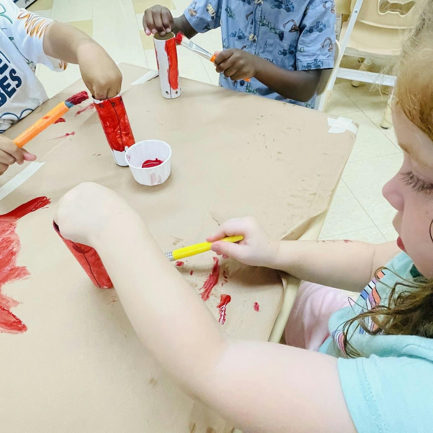 Children painting cardboard tubes with red paint at a table