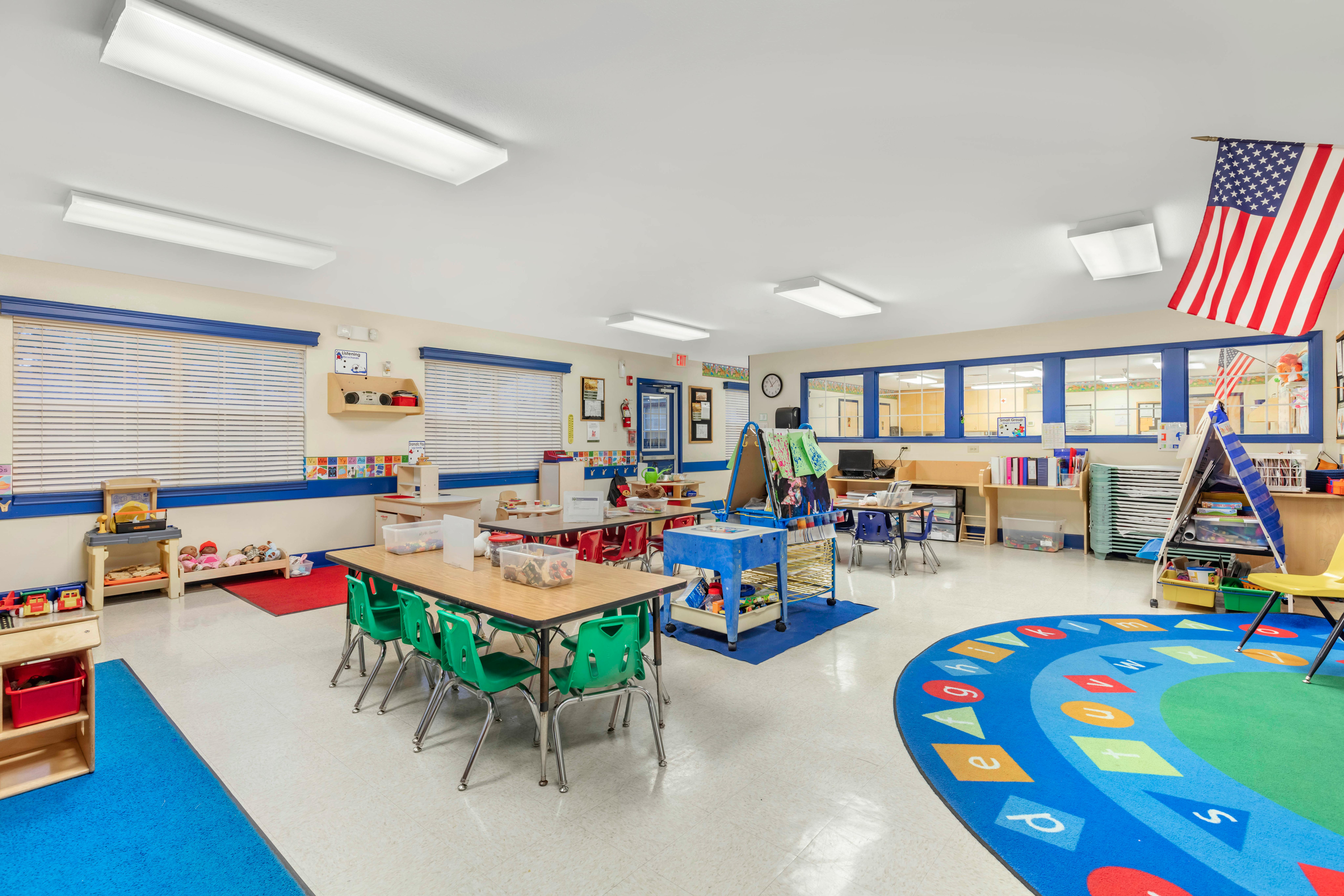 A colorful classroom with tables and chairs for children, activity centers, and a flag