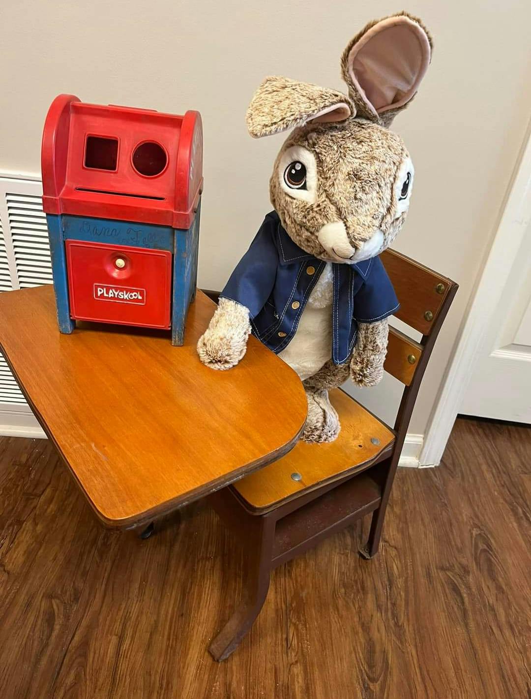 Stuffed rabbit in a denim jacket sitting at a school desk with a toy mailbox