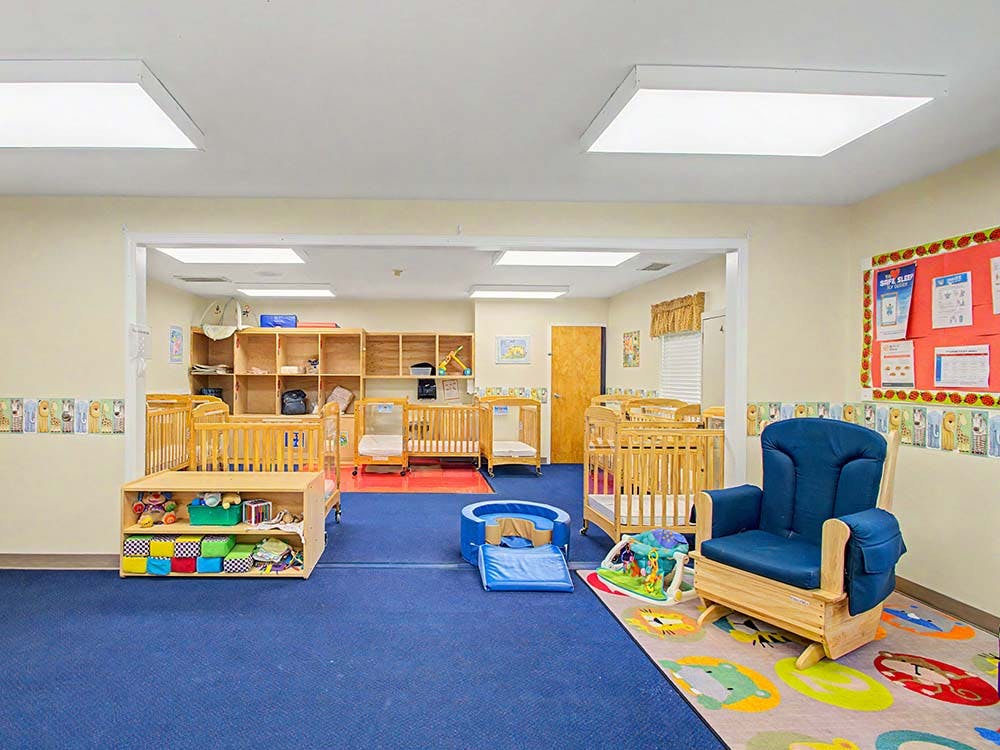 A brightly lit nursery room with multiple wooden cribs and a blue rocking chair