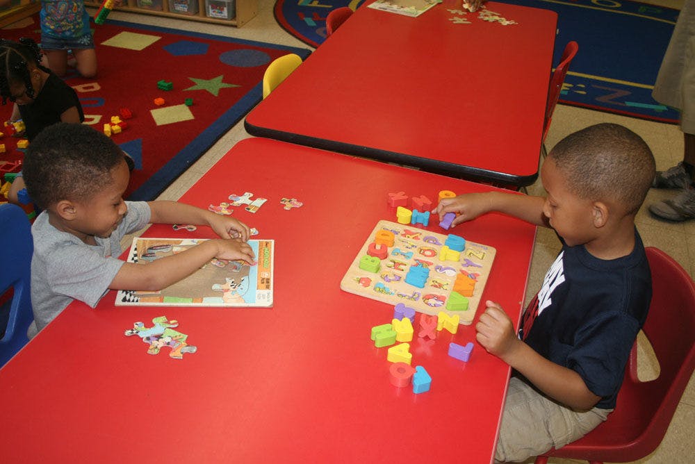Two children playing with puzzles at a red table in a classroom