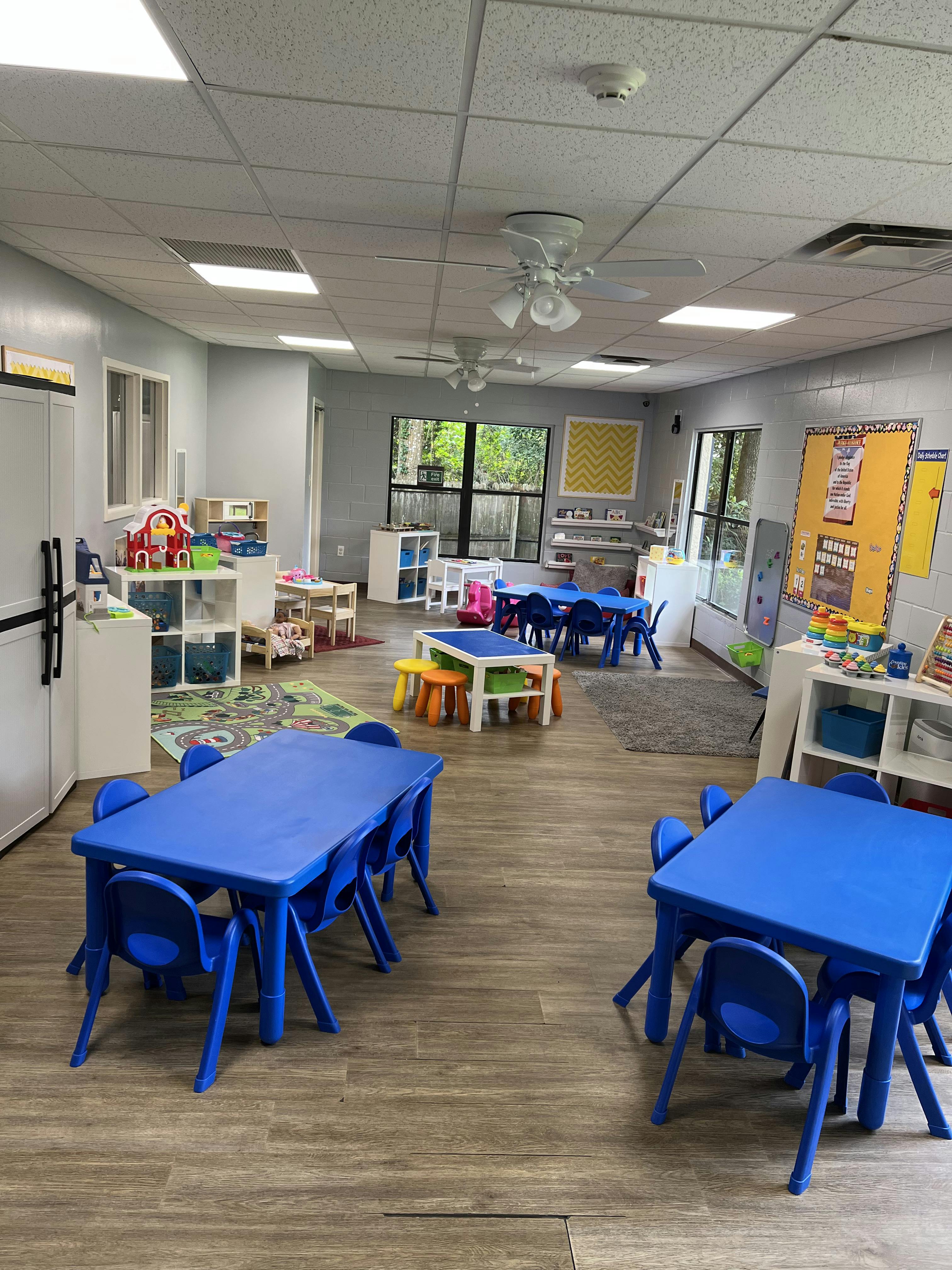 Classroom with blue tables and chairs, play areas, and a visible "Daily Schedule Chart"