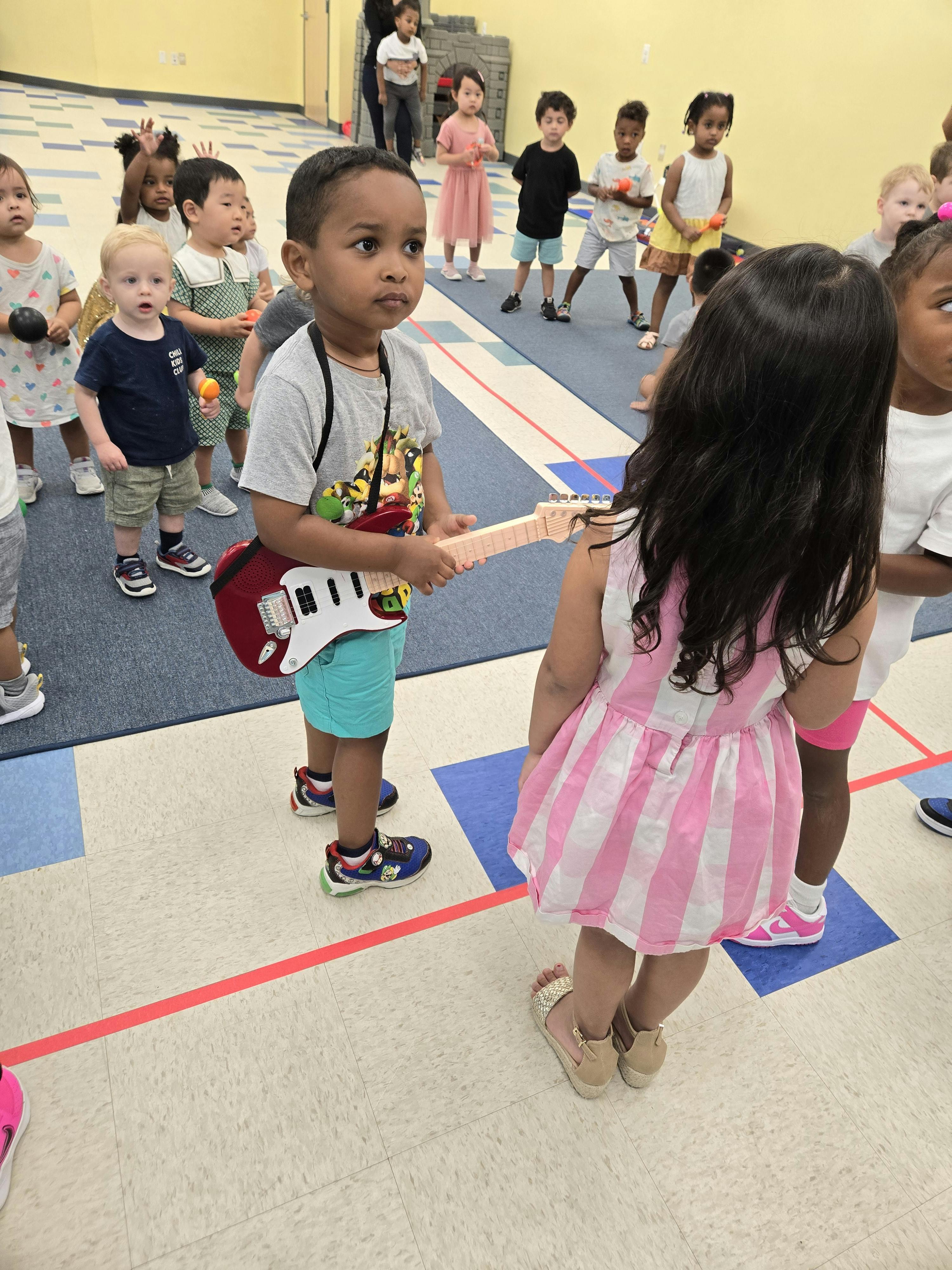Child holding a toy guitar in a classroom with other children