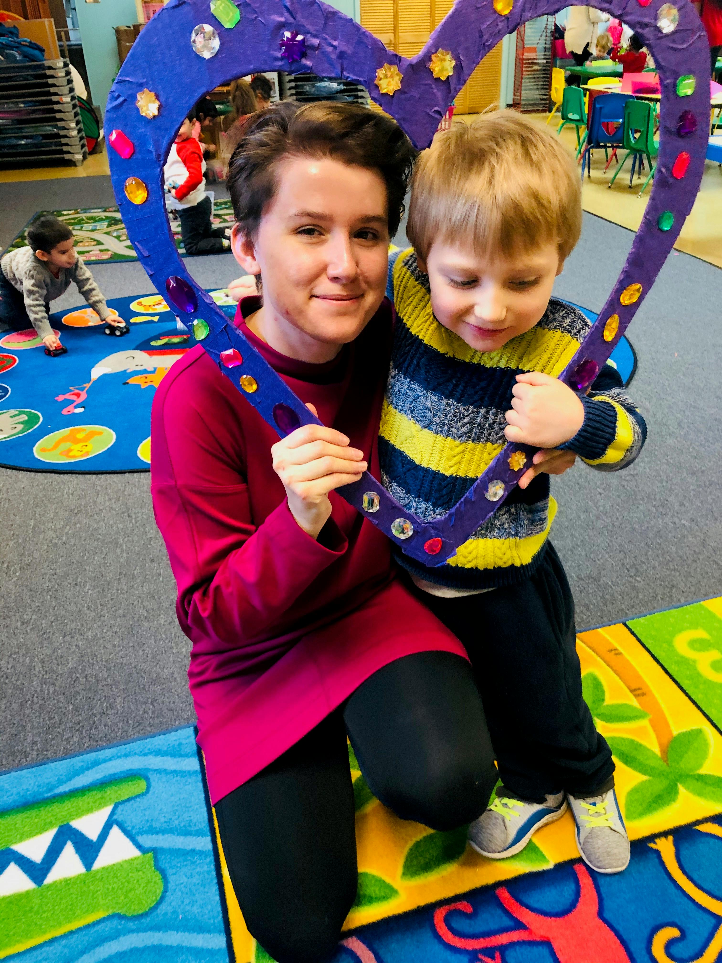 Person and child holding a heart-shaped frame in a classroom