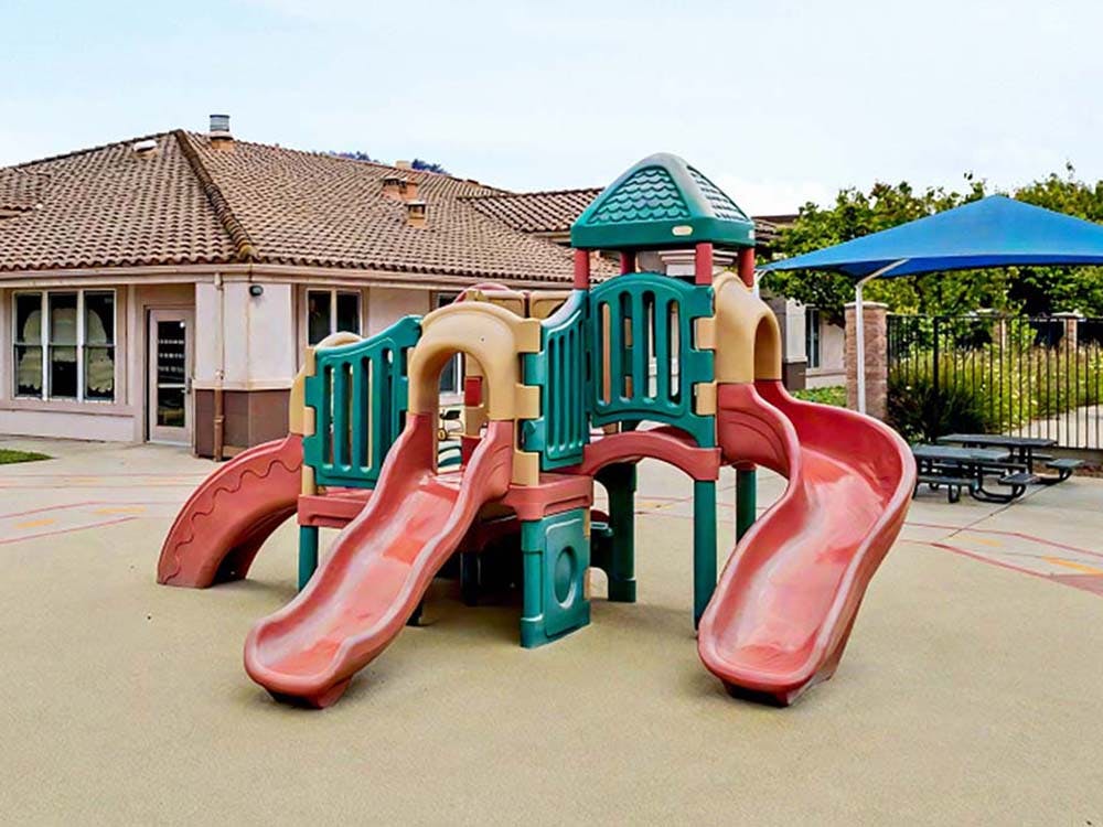 A multi-slide playground structure with a building and blue umbrella in the background