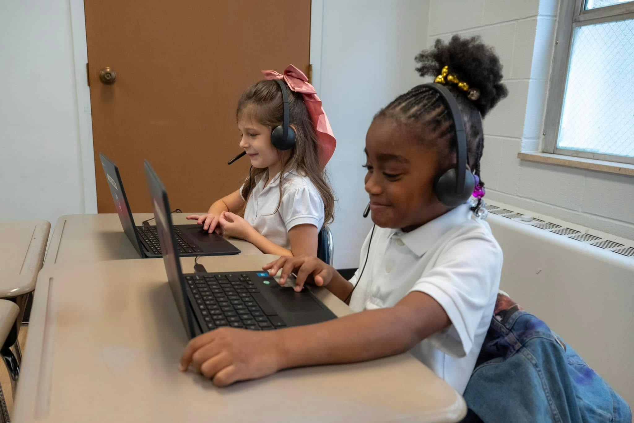 Two children wearing headsets using laptops at desks in a classroom