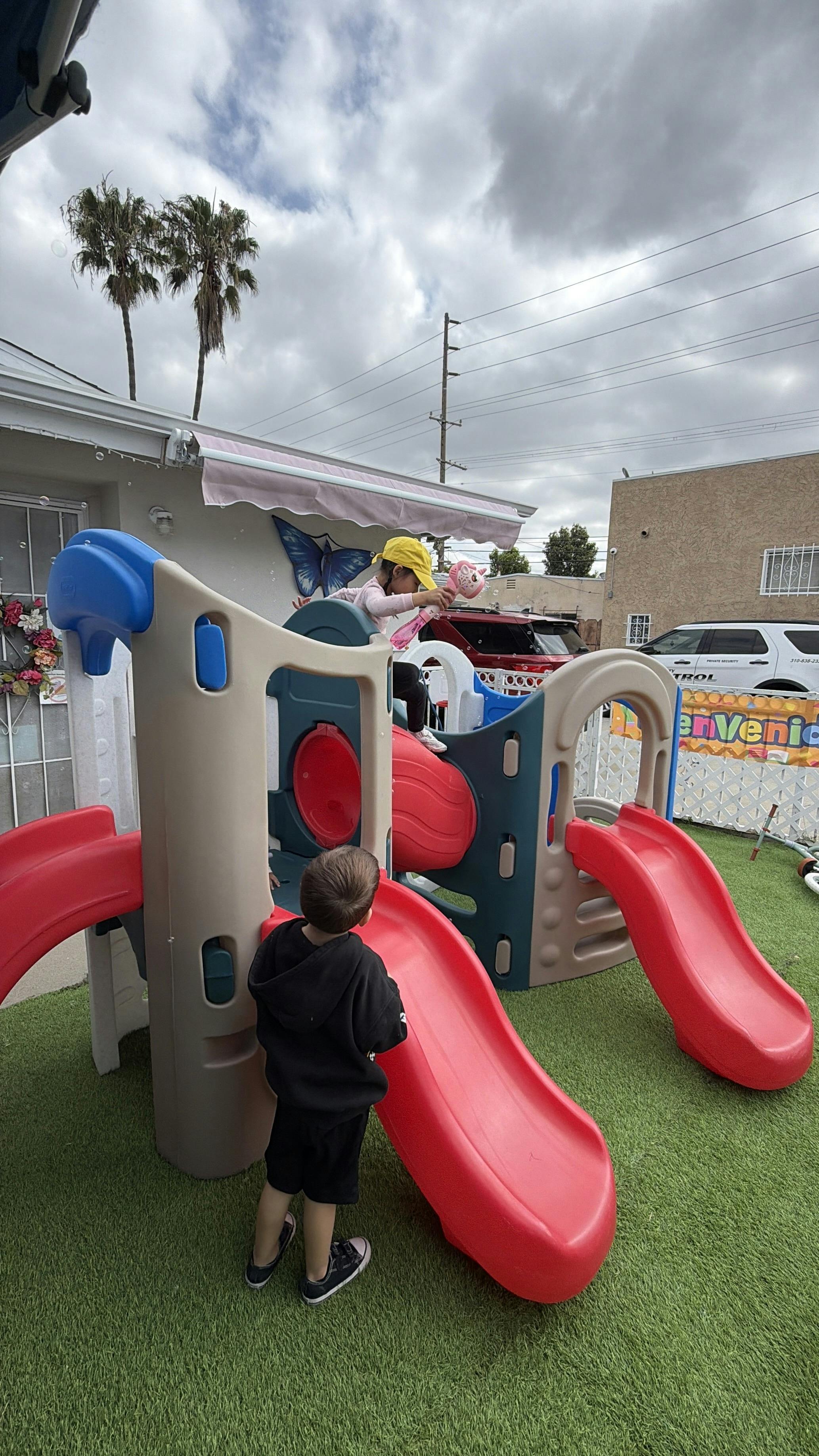 Child playing on a red and beige slide structure, another child stands below