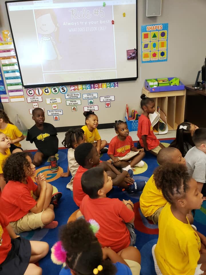 Children sitting on a rug in a classroom looking at a screen