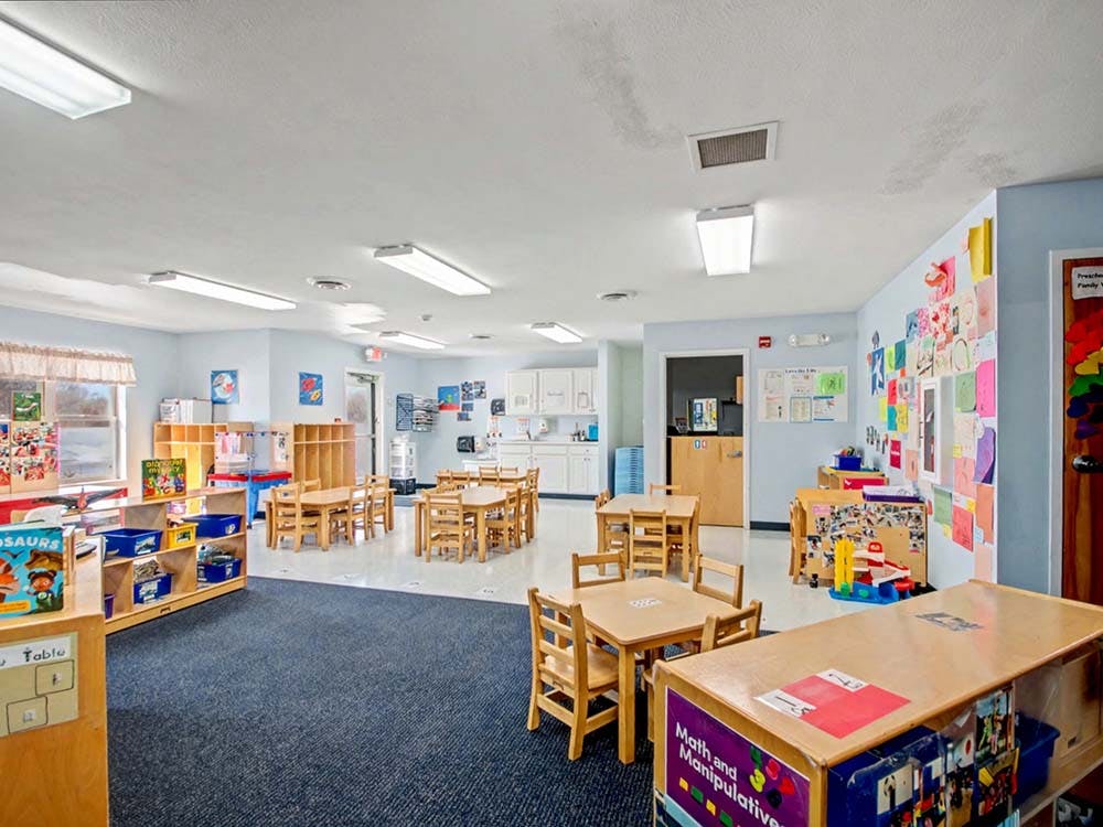 Classroom with small wooden tables and chairs, shelves, and artwork on walls