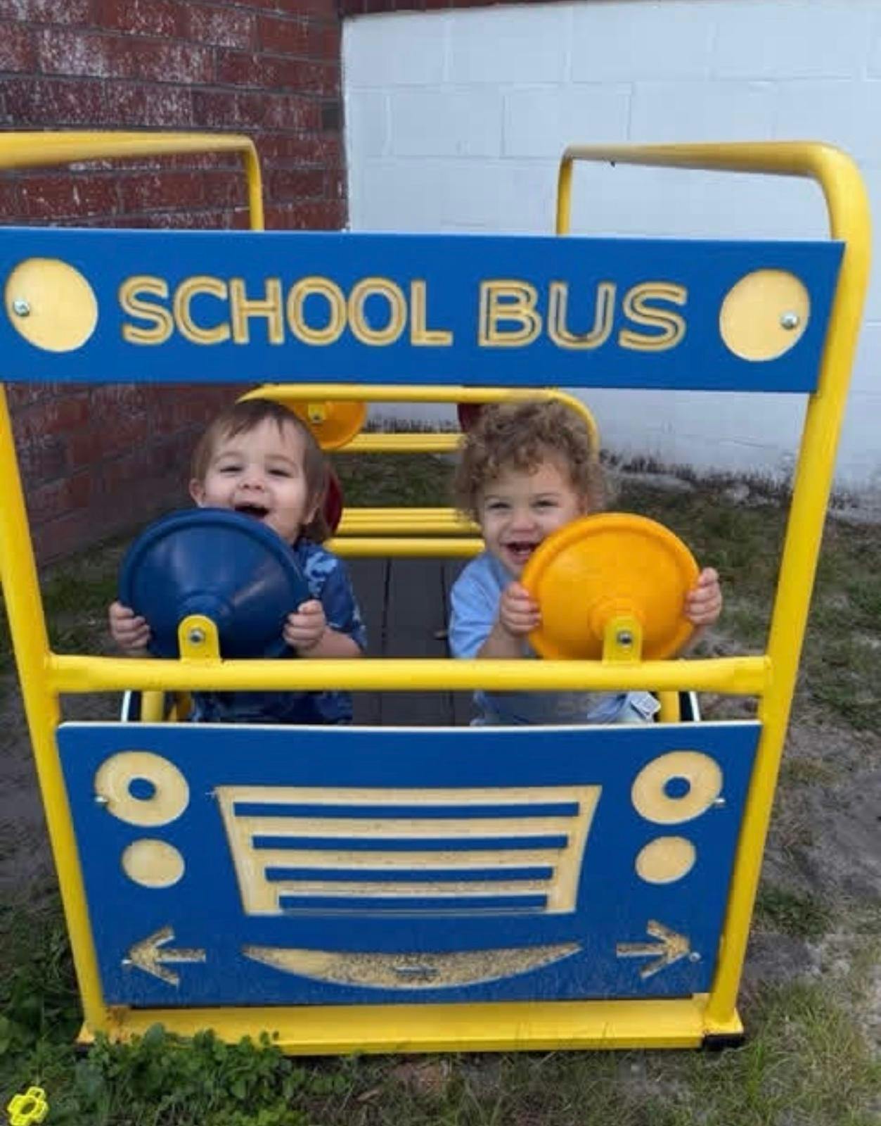 Two children sitting in a school bus playground structure holding steering wheels