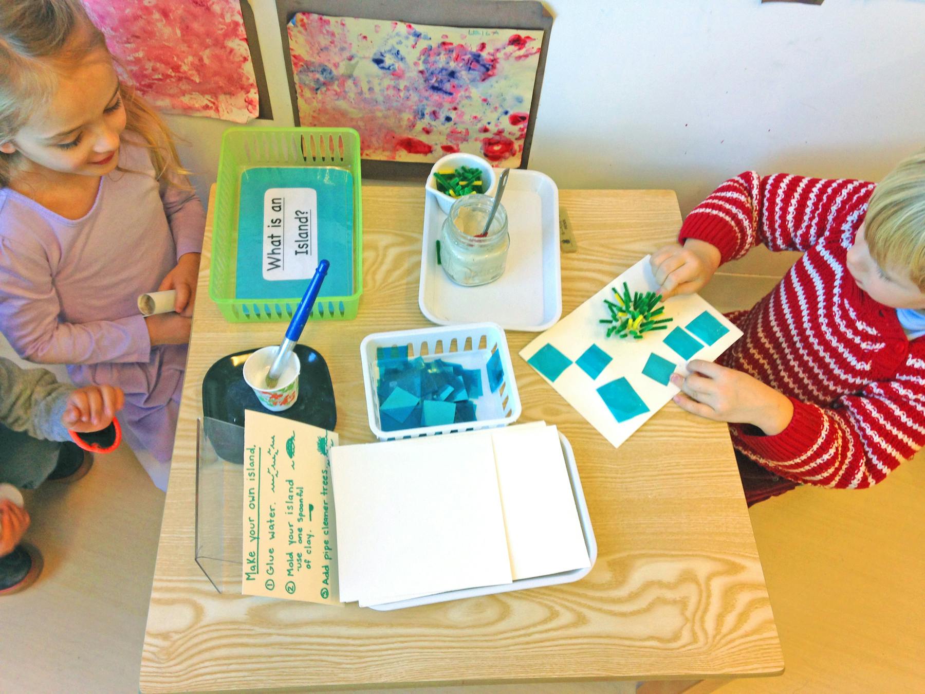 Two children at a table with craft supplies, one working on a paper craft