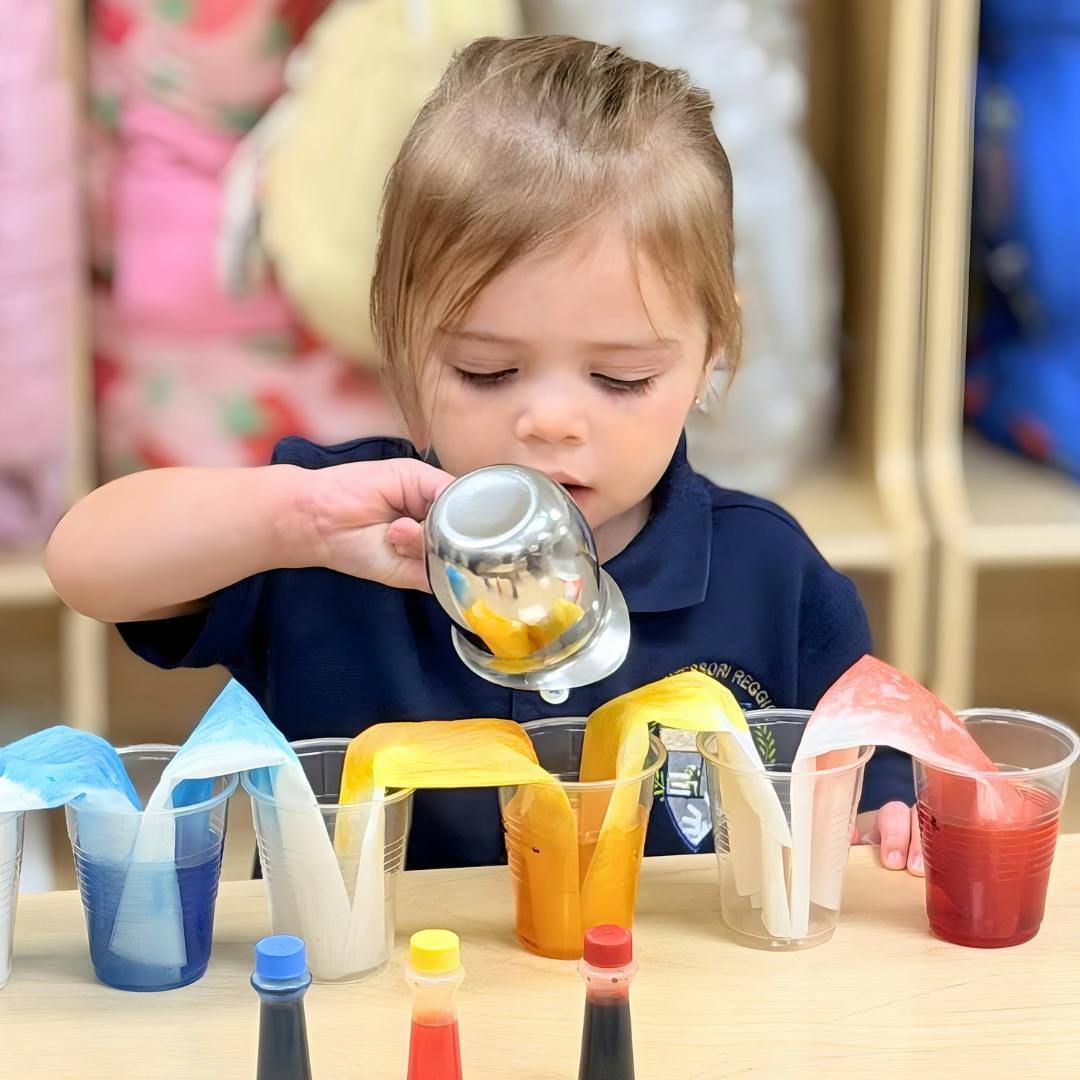 Child pouring liquid from a small metal cup into clear plastic cups with colored liquid