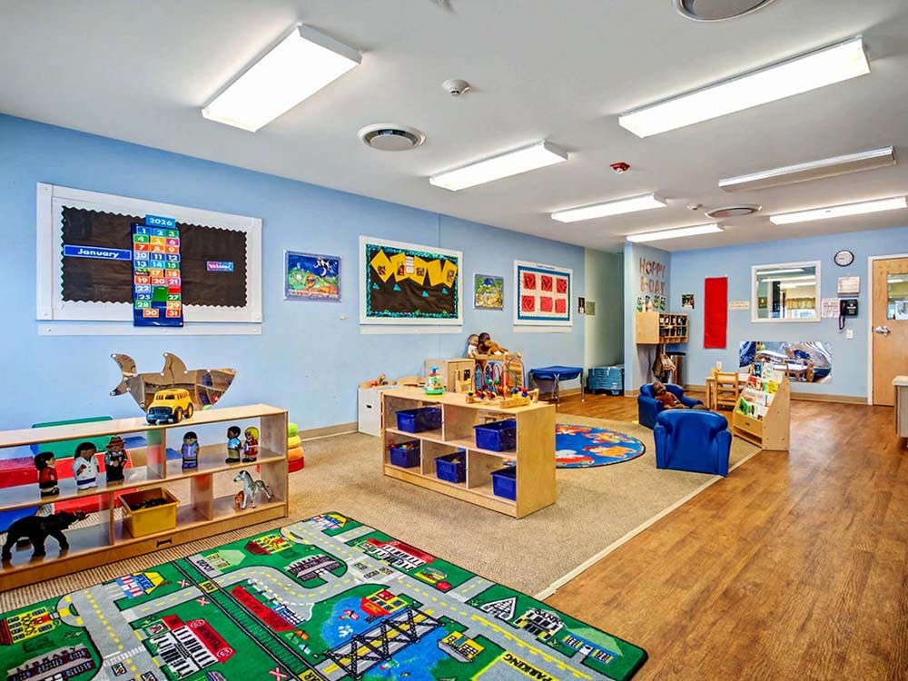 A colorful classroom with play mats, low shelves of toys, and small chairs