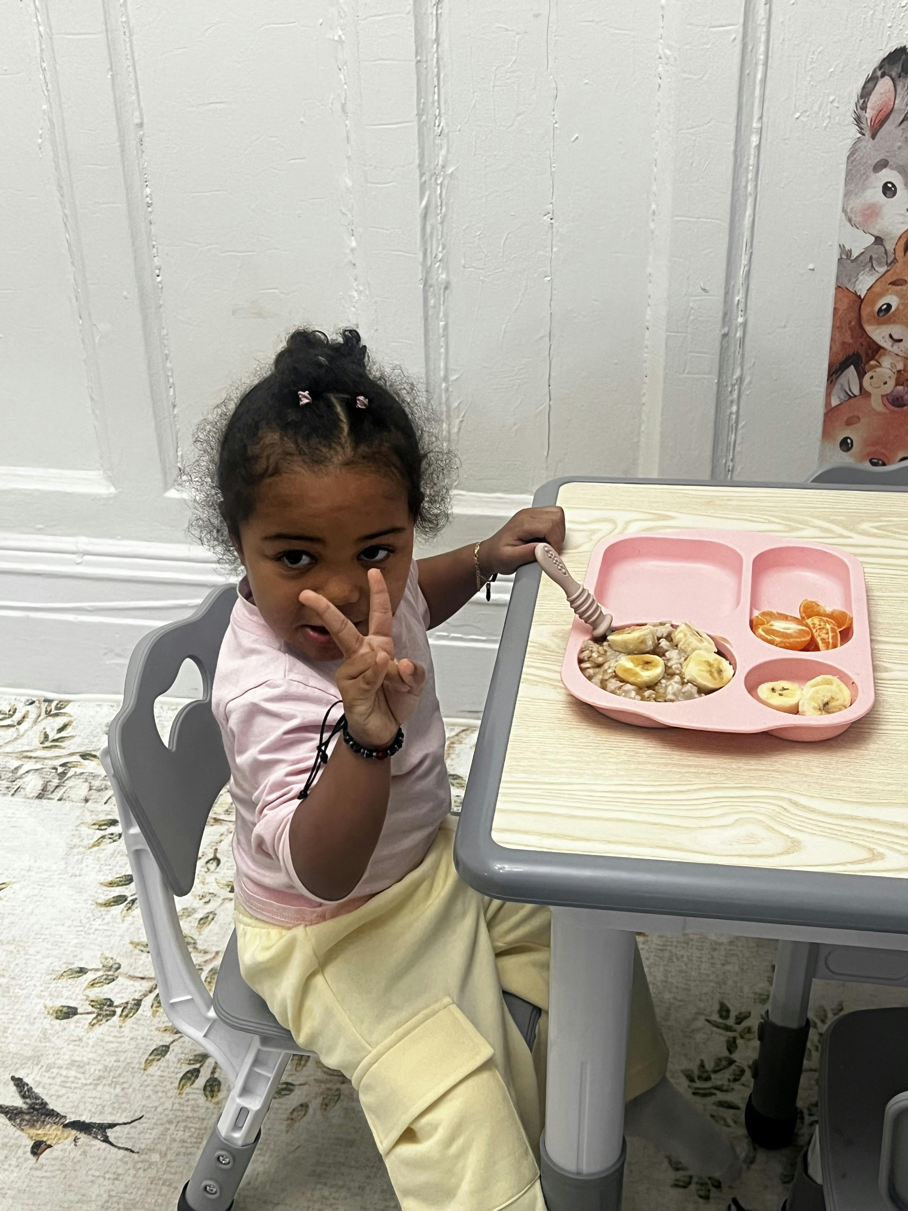 Child sitting at a table with a pink plate of food, holding up two fingers