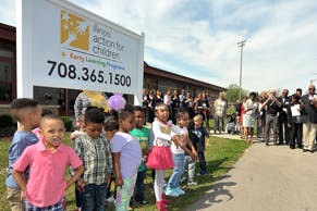 Children standing in front of a building with a sign, adults in background